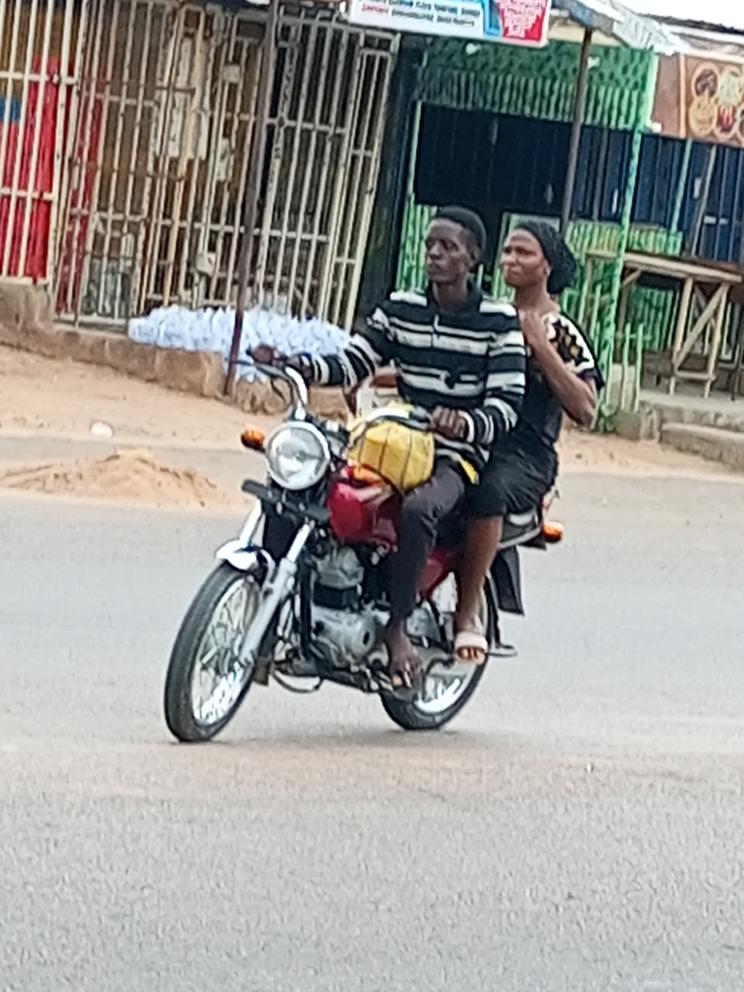 Two individuals, a male and a female, are riding on a red motorcycle on a paved road. The male is driving, wearing a striped long-sleeved shirt and dark pants. The female is a passenger behind him, wearing a headscarf and a patterned top. They are both looking forward. The motorcycle has a yellow bag strapped to the back and is equipped with a headlight and mirrors. The background shows a street scene with buildings, some appearing to be shops, and metal grates. There are stacks of white plastic bottles on the ground to the left of the motorcycle. The scene is likely in Gombe, Nigeria, a city known for its commercial activities and bustling street life, with similar architecture and road conditions often found there.