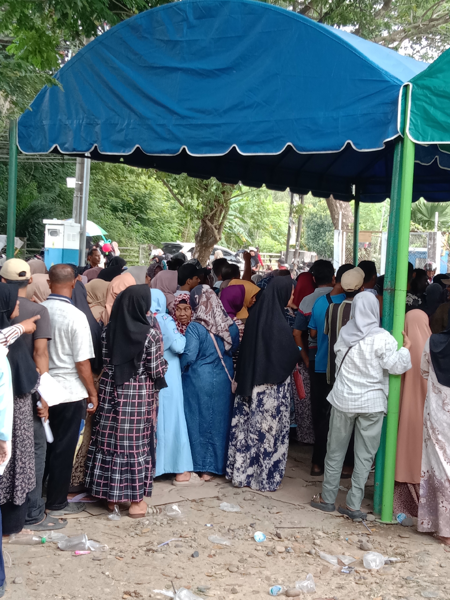 A crowd of people stands under a blue and green awning in Lhokseumawe, Indonesia. Most of the people are wearing hijabs. The ground is littered with trash. Some people are holding white pieces of paper.