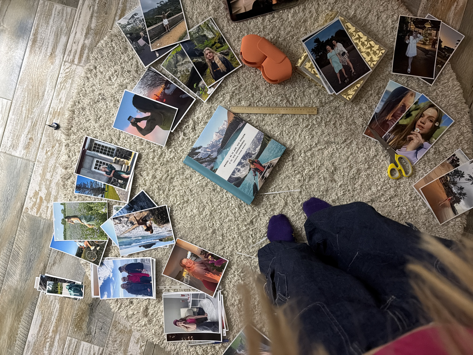 A high-angle indoor shot shows an arrangement of photos and objects on a beige shag rug, situated between gray wood-look floor tiles and the legs of an individual. The person, wearing denim jeans and purple socks, is positioned in the lower right of the frame.
Scattered across the rug are numerous printed photographs depicting various subjects, including landscapes, portraits of people, and travel scenes. Central to the arrangement is a teal-colored book with Russian text printed on its cover. A wooden ruler and a pair of orange headphones lie nearby. A pair of yellow-handled scissors and a small tube of glue are also present on the rug.
The flooring transitions from the gray, wood-look tiles on the left side of the image to the rug in the center, creating a distinct separation of space.
