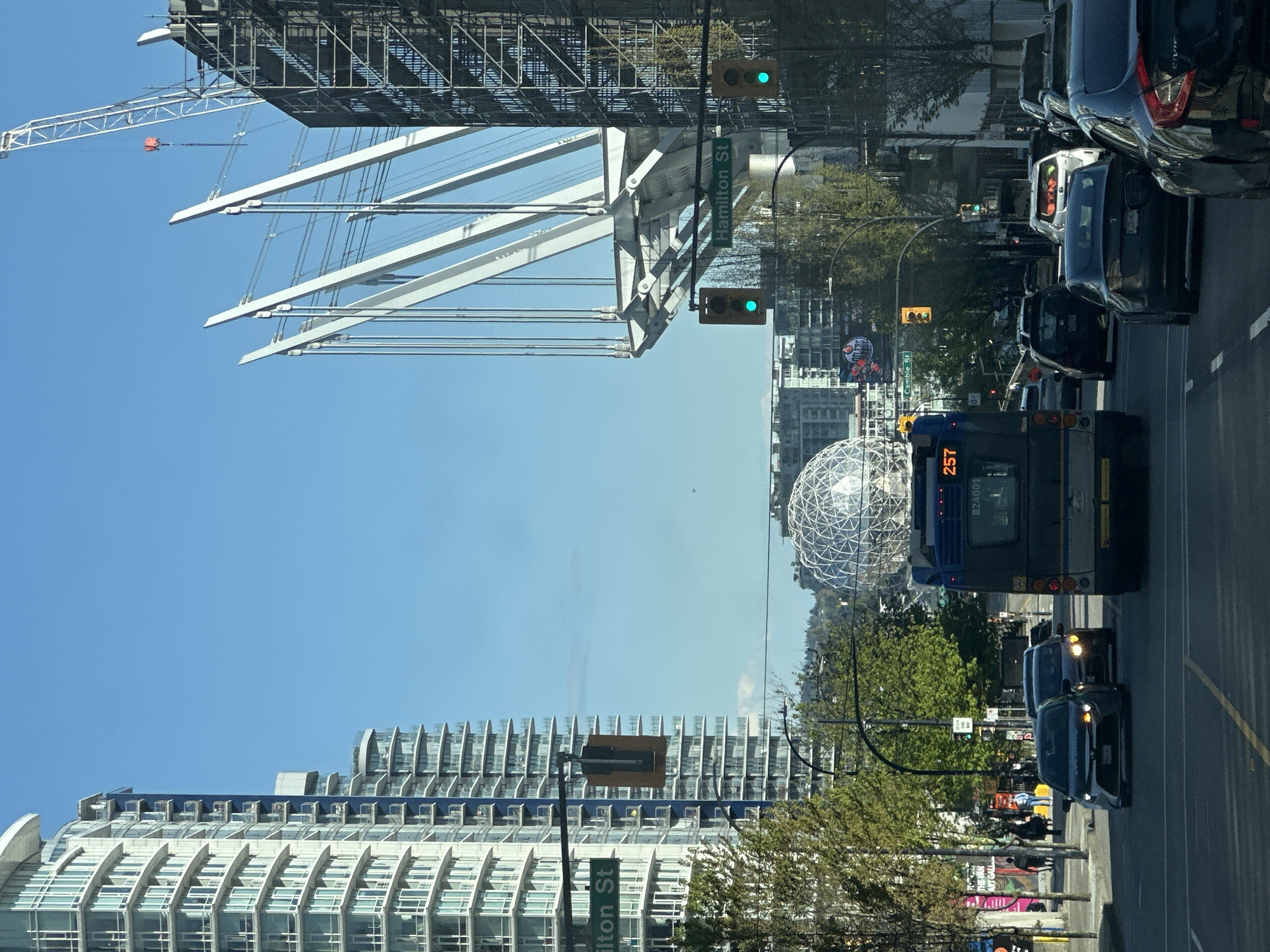 The image shows a street in Vancouver, Canada. On the right side of the street, there are several cars and a bus numbered 257 in a traffic jam. On the left, there are several tall buildings, including BC Place Stadium and Science World. The sky is clear and blue. The street sign shows "Hamilton St."