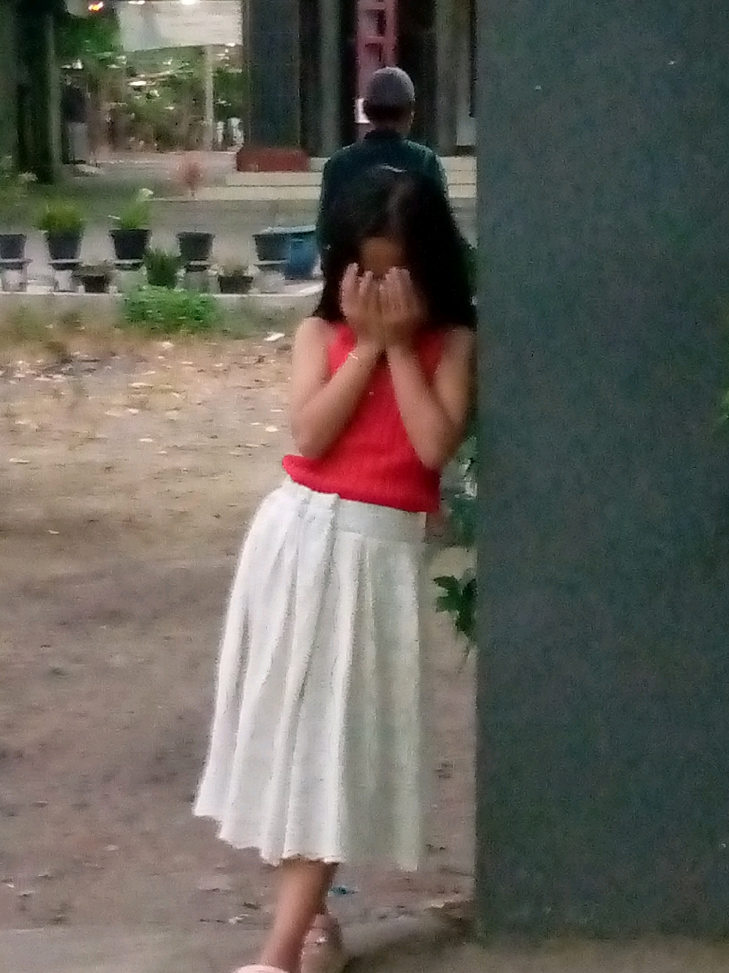 In Sumberwono, Indonesia, an out-of-focus image captures a young girl with dark hair, wearing a red tank top and a white pleated skirt, standing in front of a dark gray, textured wall. She is covering her face with her hands, suggesting she might be embarrassed or crying. Behind her, another person, possibly an adult, is visible from the shoulders up, wearing a dark cap and a dark jacket, facing away from the camera. The ground appears to be dirt or gravel, with some scattered leaves. In the background, a row of potted plants is visible, some with green foliage. The lighting suggests it is daytime, possibly overcast.