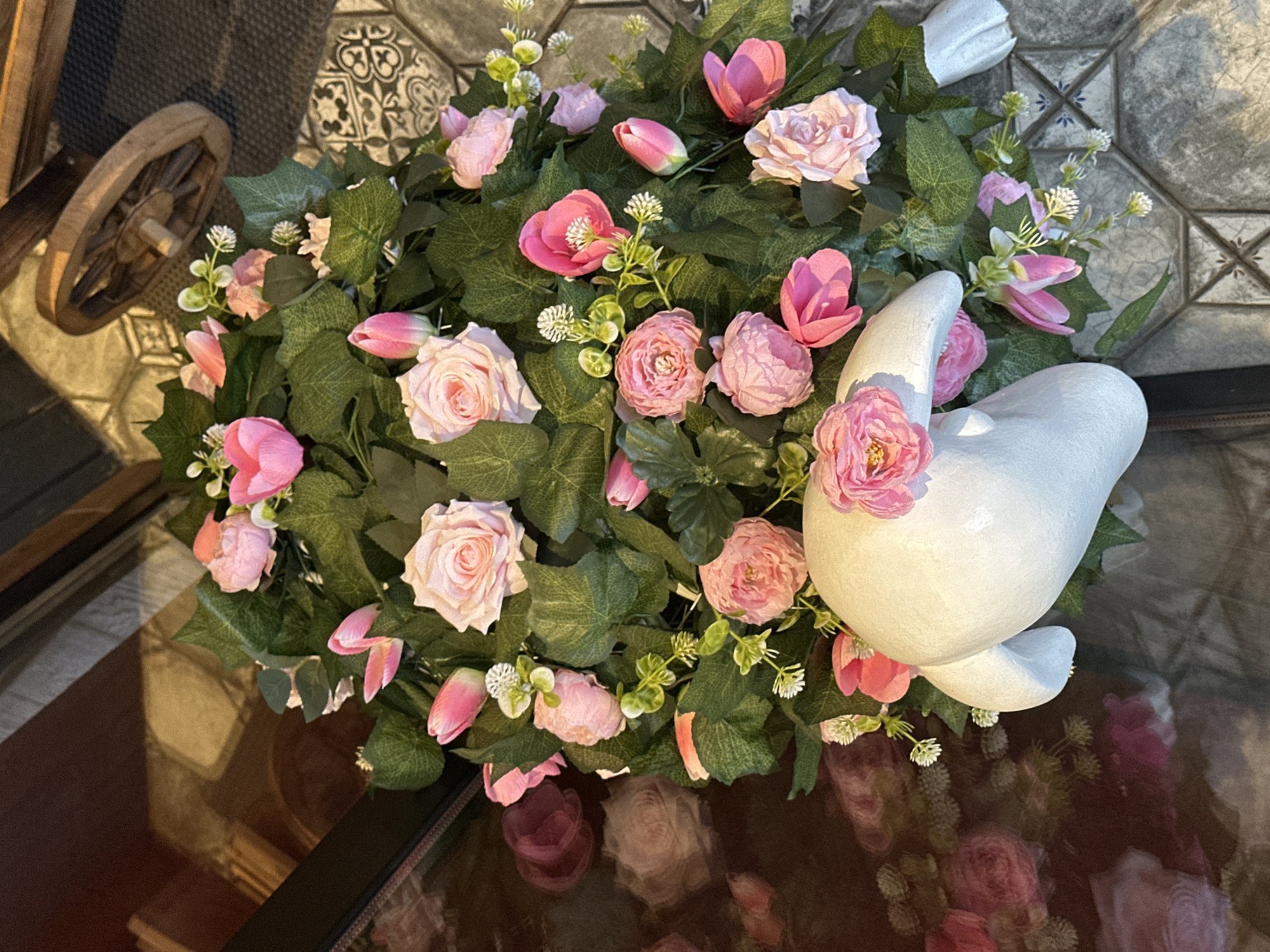 A top-down view shows an arrangement of pink roses, tulips, and green foliage encircling a white, stylized bird statue. The arrangement sits atop a shiny, reflective surface, likely a glass table, which mirrors the flowers and statue, creating a blurred reflection of them. To the left, a partial view of a wooden wheel suggests a decorative element within the scene. The background includes patterned tiles in shades of grey and white. The location, Izhevsk, Russia, cannot be verified from the provided visual information. There is no information about people, interactions, or specific actions.
