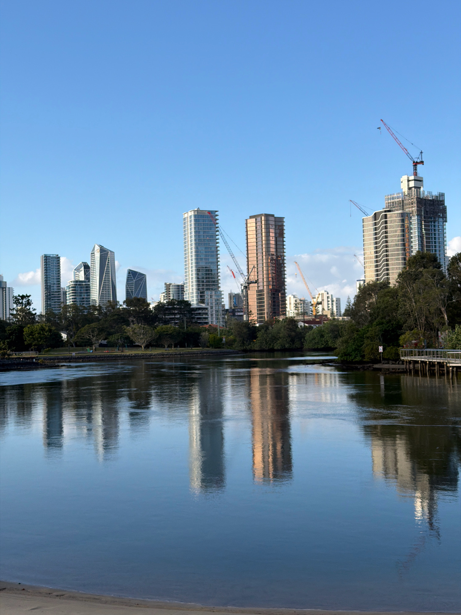 The image captures a serene waterfront view of the Broadbeach Waters skyline in Australia. The foreground is dominated by a calm, reflective body of water, likely the Nerang River or a canal, with a sandy shore visible at the bottom. The water mirrors the impressive cityscape that rises behind a line of lush green trees.
The skyline is characterized by a collection of modern high-rise buildings, including several sleek skyscrapers and apartment complexes. Some of these structures appear to be under construction, indicated by the presence of cranes and unfinished sections. One particularly striking building features a distinctive geometric, faceted design. Another prominent building exhibits a warm, coppery hue.
The sky is a clear, vibrant blue, with a few wispy white clouds scattered across it. The lighting suggests it is either early morning or late afternoon, casting soft light and creating reflections on the water. The environment appears clean and well-maintained, with a boardwalk or pier visible on the right side of the frame, hinting at public access to the waterfront. No people are distinctly visible in the image, contributing to a sense of peaceful tranquility.