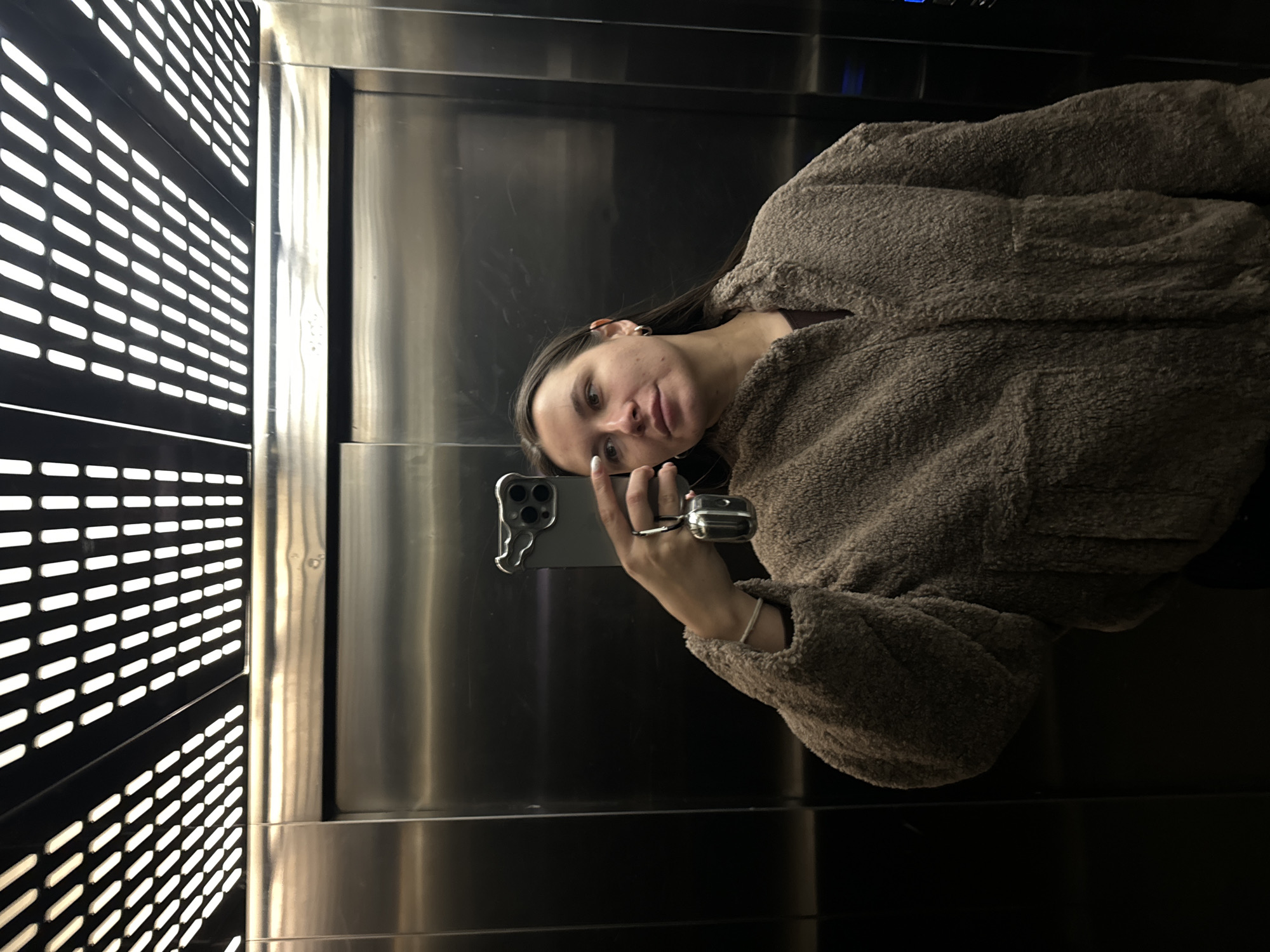 A young woman is taking a selfie in what appears to be an elevator. She is wearing a brown, plush jacket and is holding a smartphone in her left hand. The elevator's interior features a wall with a grid-like pattern of horizontal slits and a metallic wall with reflective surfaces. The photograph was taken in Odintsovo, Russia.