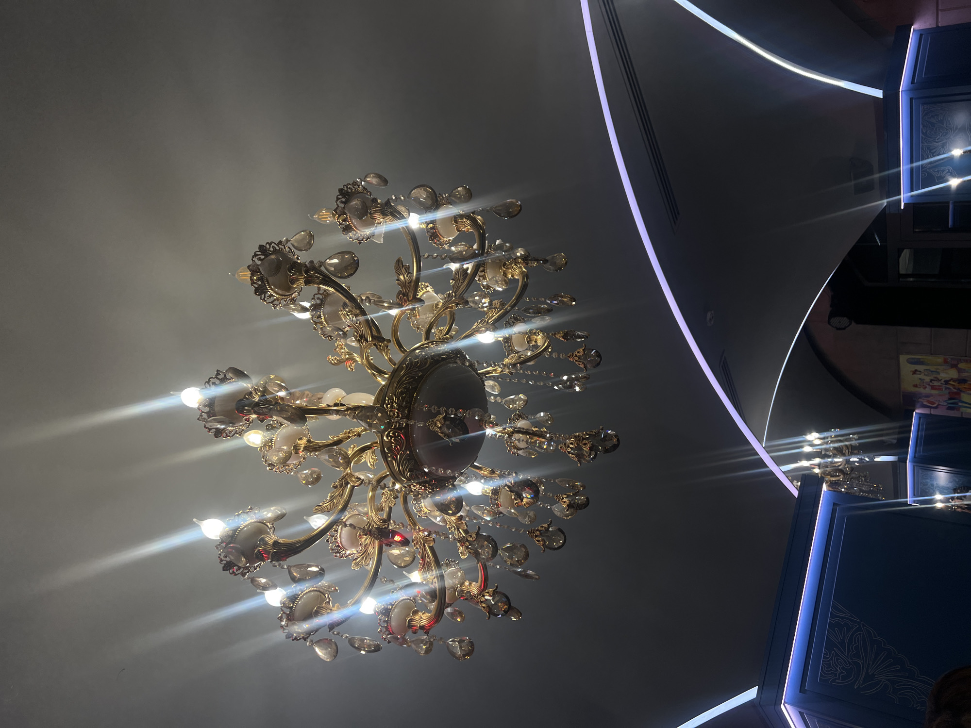 The image depicts a ceiling with an ornate chandelier, situated in an interior location in Yerevan, Armenia. The chandelier features multiple light sources, likely incandescent bulbs, emitting bright light and causing light reflection on its crystal and gold-colored components. The ceiling is painted a light gray color. To the right of the chandelier, parts of architectural elements are visible, with blue and white neon lighting. These appear to be decorative panels or structures within the building. The overall ambiance is modern. A small portion of a person's head is visible in the lower right corner.