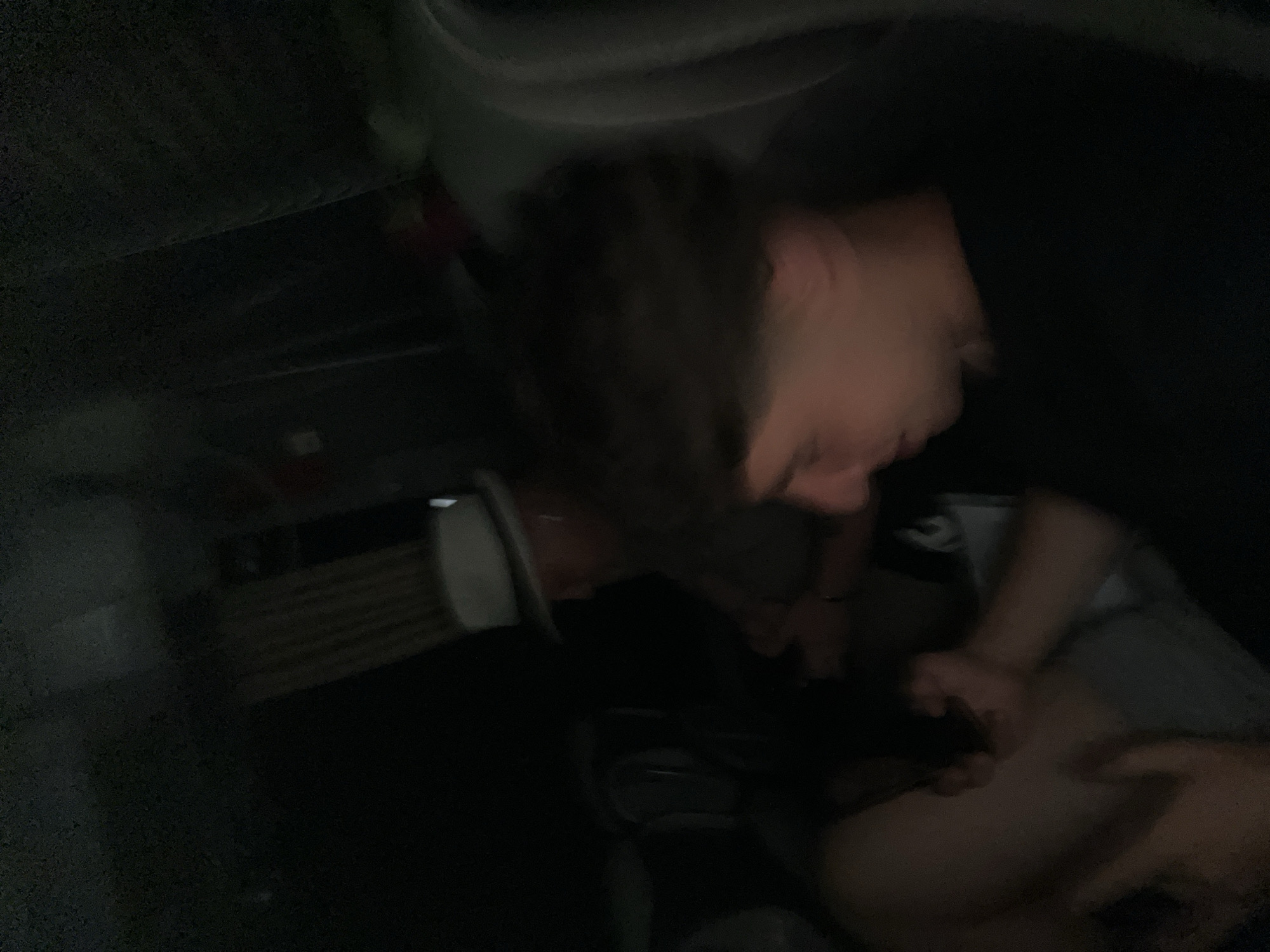 A blurred, dark, indoor photograph shows a person with short hair wearing a black shirt, leaning over and appearing to interact with someone whose legs are visible. An unidentified object is in the background. The location is unknown.