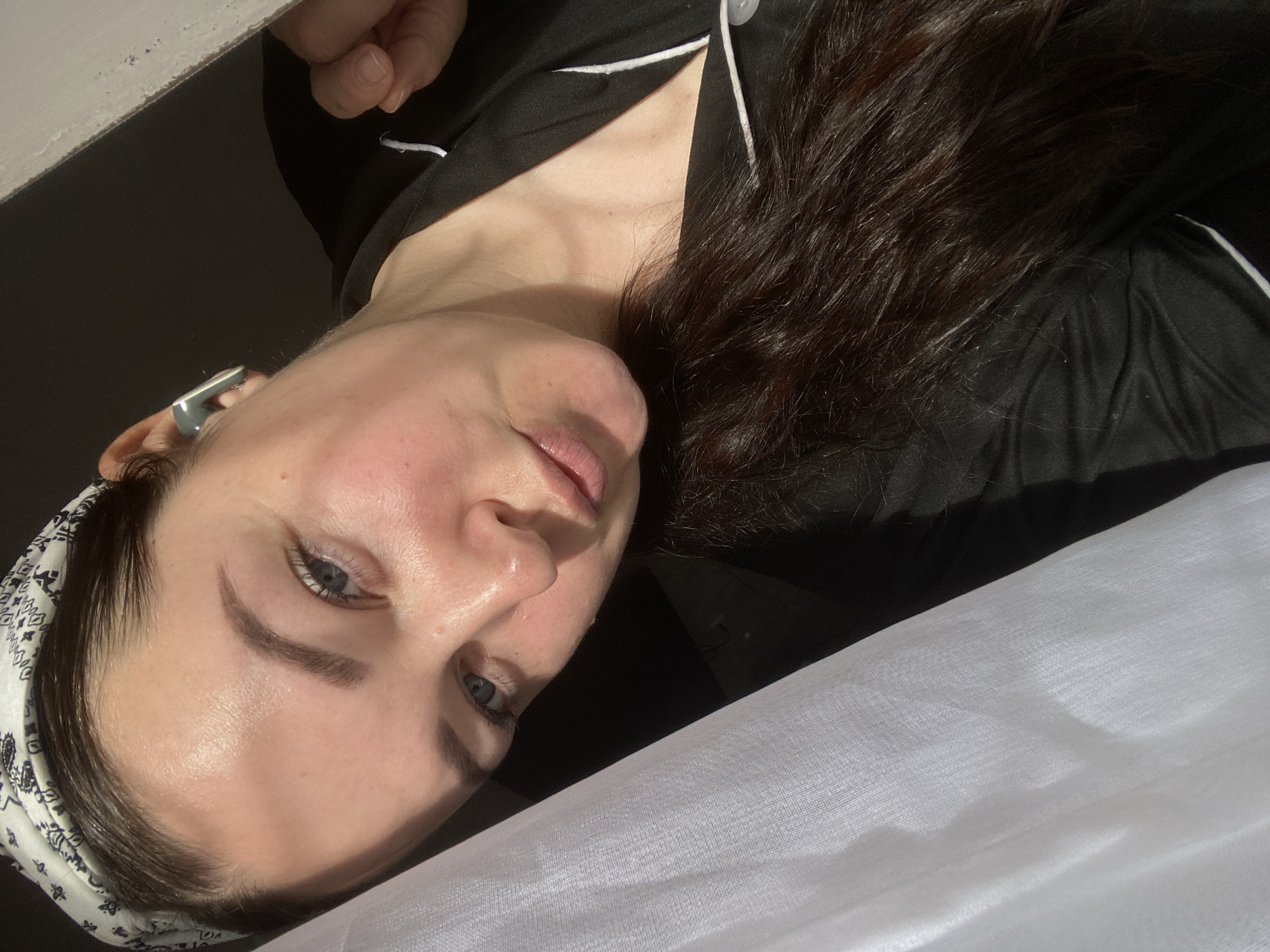 A medium close-up shot depicts a fair-skinned woman with dark brown hair and blue eyes, angled to the right. She wears a black shirt with white piping and a paisley-patterned headband. An electronic device is visible in her ear. The background consists of a dark area above, a white, gauzy fabric below, and a possible window or wall in the upper left corner. There is no information in the picture to suggest the location is Sochi, Russia.