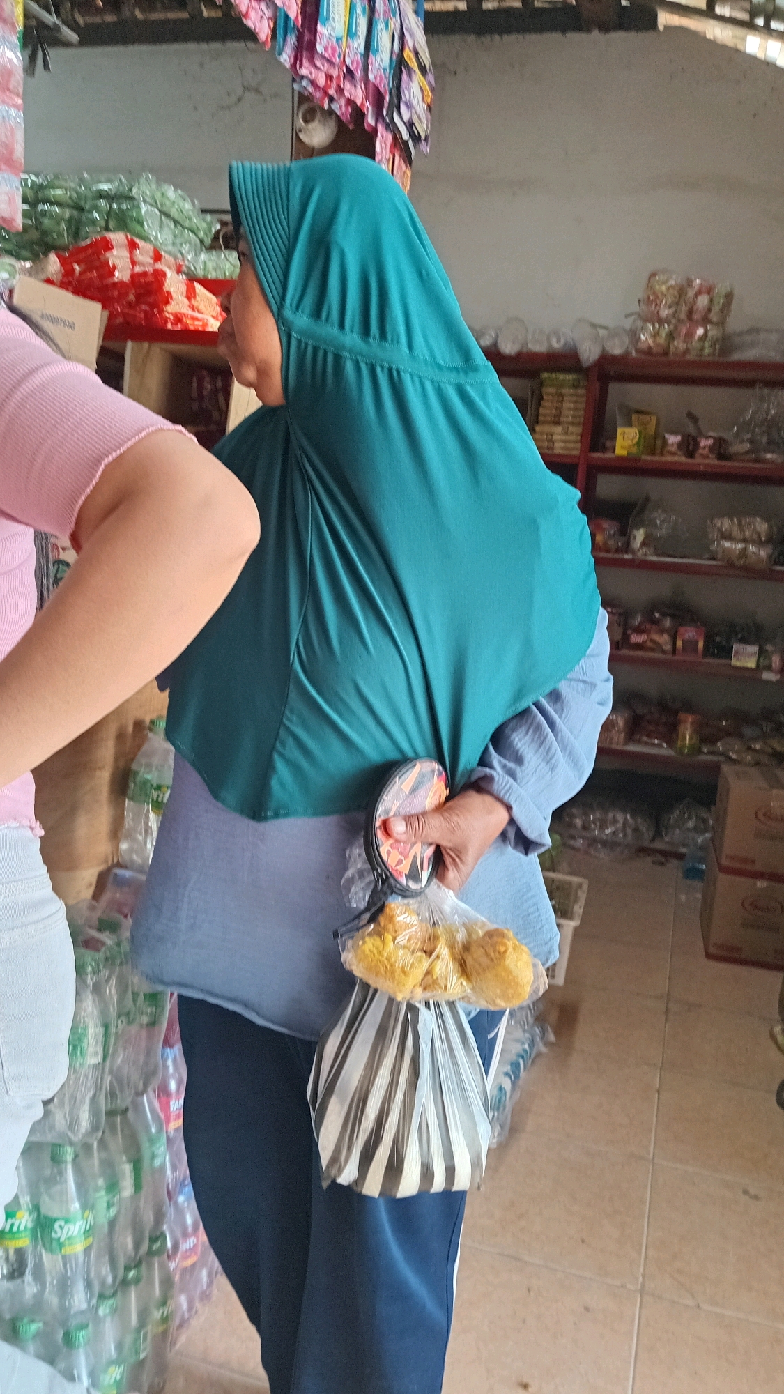 In Kedungwaru, Indonesia, a woman wearing a teal hijab and a light blue blouse is standing in a shop. She is holding a circular object with a patterned design in her right hand, and a plastic bag filled with yellow, fried food items in her left hand. Another person, wearing a pink top and white pants, is partially visible on the left side of the frame. The shop appears to be stocked with various goods, including bottles of Sprite on the left, packaged items on shelves in the background, and boxes stacked on the right. The floor is tiled.