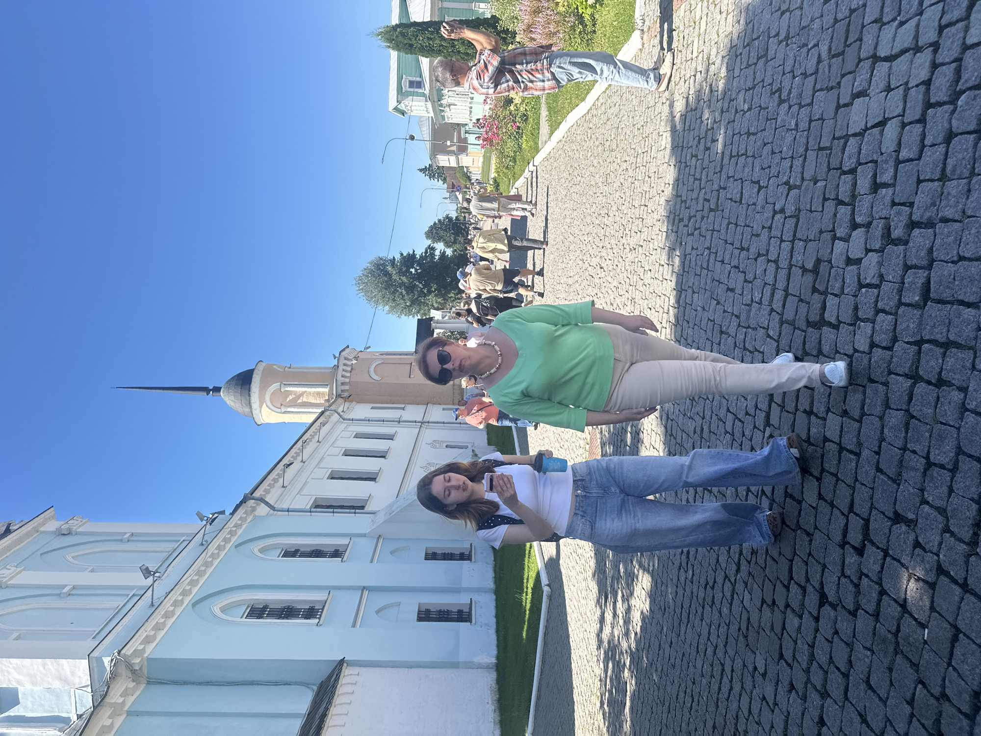 In Kolomna, Russia, on a bright, sunny day, two women walk on a cobblestone pathway in front of a light blue building. The building has arched windows with dark bars and a white decorative trim. Further down the path, a man in a plaid shirt and jeans takes a photo with his phone, and other people stroll along the path, some with flowers in the foreground. The sky is clear blue, and the overall scene appears to be a historic or religious site.