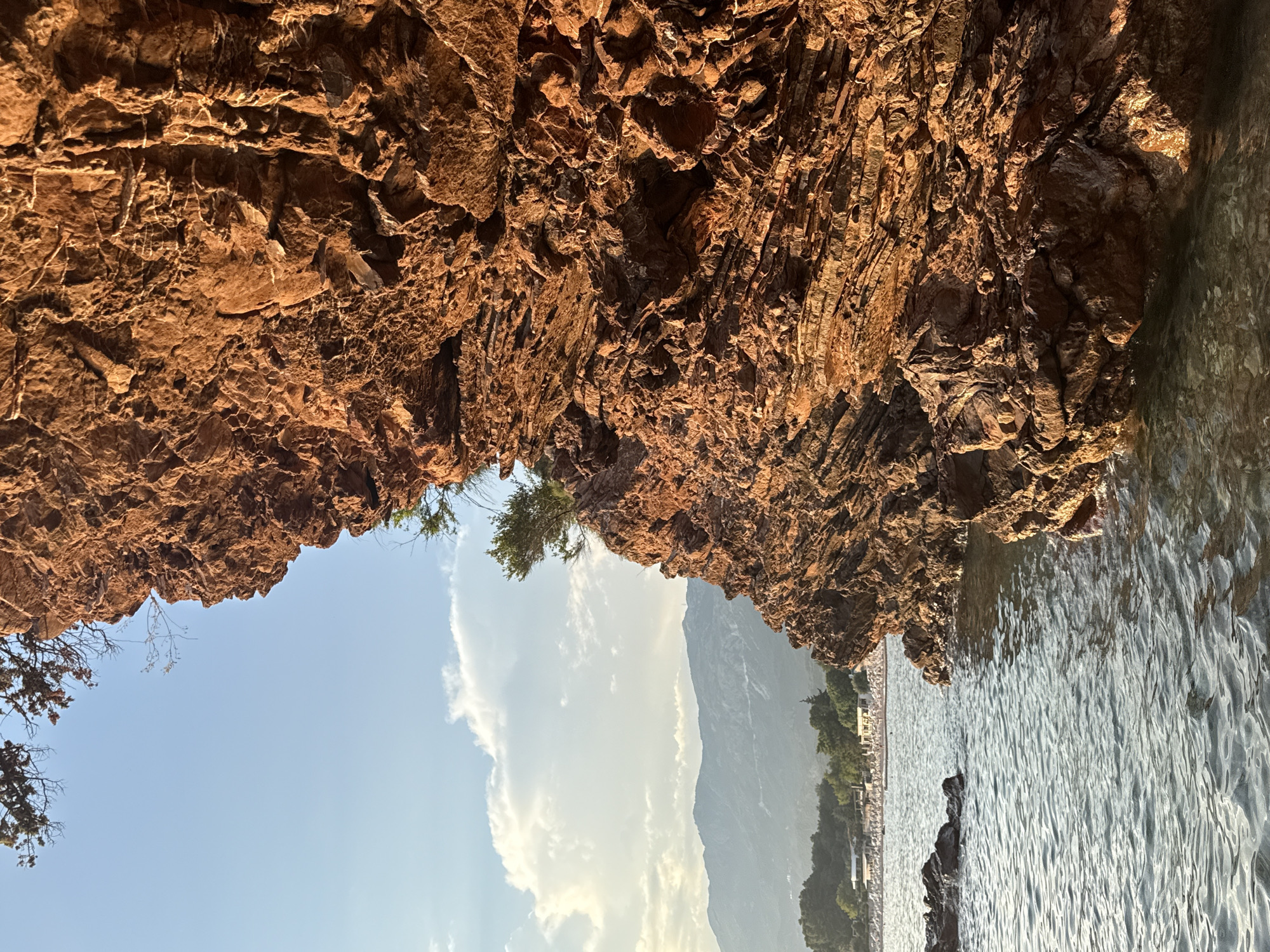 The image shows a dramatic view of a rocky coastline with the sea to the right and the sky above. The rocks are a warm, reddish-brown color and have a rough, layered texture. Sunlight illuminates parts of the rock face, creating highlights and shadows.
Through a natural archway formed by the rocks, a scenic vista unfolds. The sky is a clear blue with a few scattered white clouds. In the distance, a hazy mountain range can be seen, with hints of vegetation and buildings along its base, suggesting a coastal town or resort. The sea is calm with gentle ripples, reflecting the light and appearing somewhat translucent.
On the left side, some dry branches and a small green tree emerge from the rock face, adding a touch of natural life to the rugged landscape. The overall impression is one of a beautiful, natural setting, possibly a cove or a cave opening onto the sea. The lighting suggests it is either late afternoon or early morning, with the sun casting a warm glow. There is no visible text in the image.