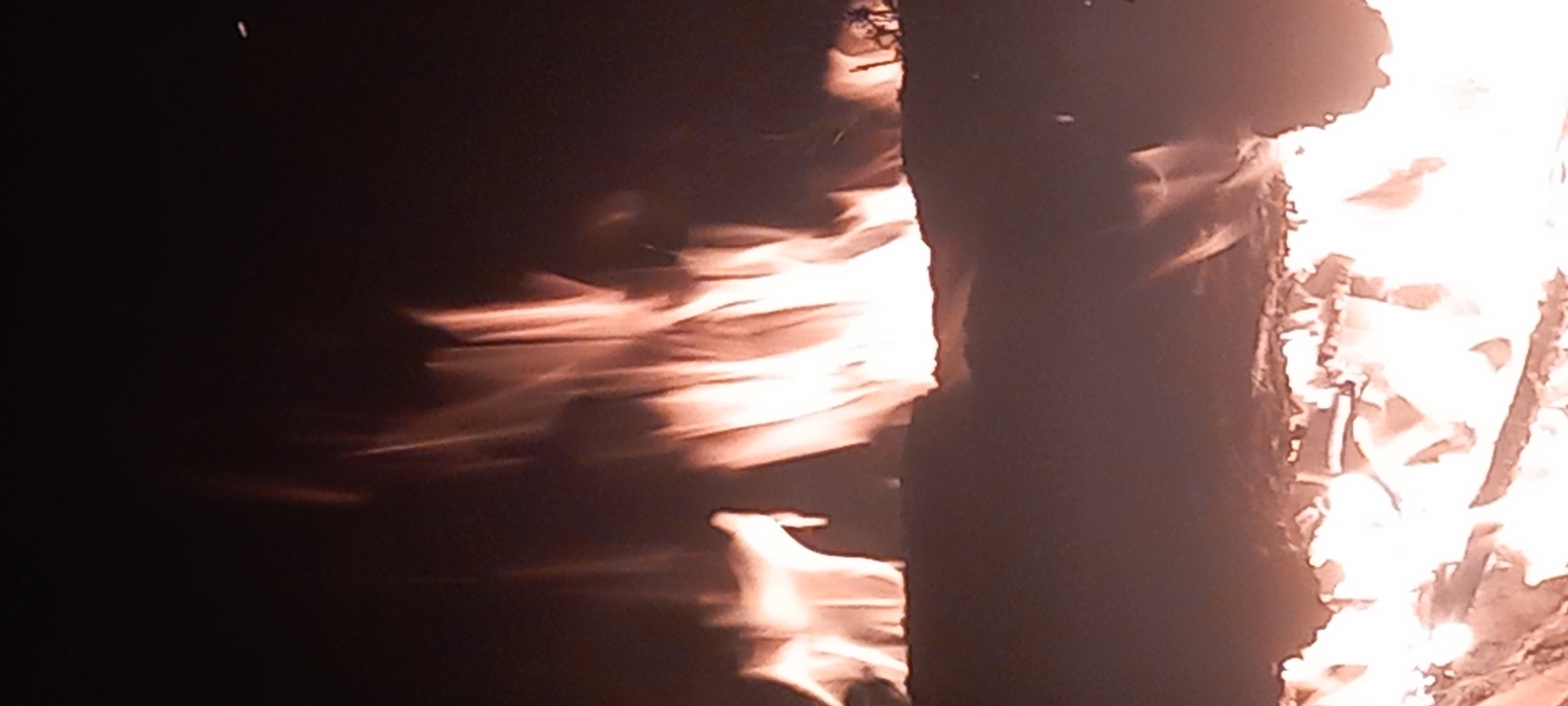 This image depicts a significant outdoor fire at night in Kryvyi Rih, Ukraine. The scene is dominated by bright flames against a dark, unlit background. The lower third of the frame shows a mass of intense, bright white and orange flames, indicating a strong combustion source. Above this, an irregular, dark horizontal structure, possibly ground or debris, divides the fire. From behind this structure, taller, more diffuse orange and red flames rise, exhibiting distinct upward-streaking patterns characteristic of burning gases and heat plumes. These upper flames are less luminous than the lower portion but still provide substantial illumination against the deep black sky. A single, small, bright spark or ember is visible in the upper right quadrant. No individuals or structures beyond the burning material are identifiable.
