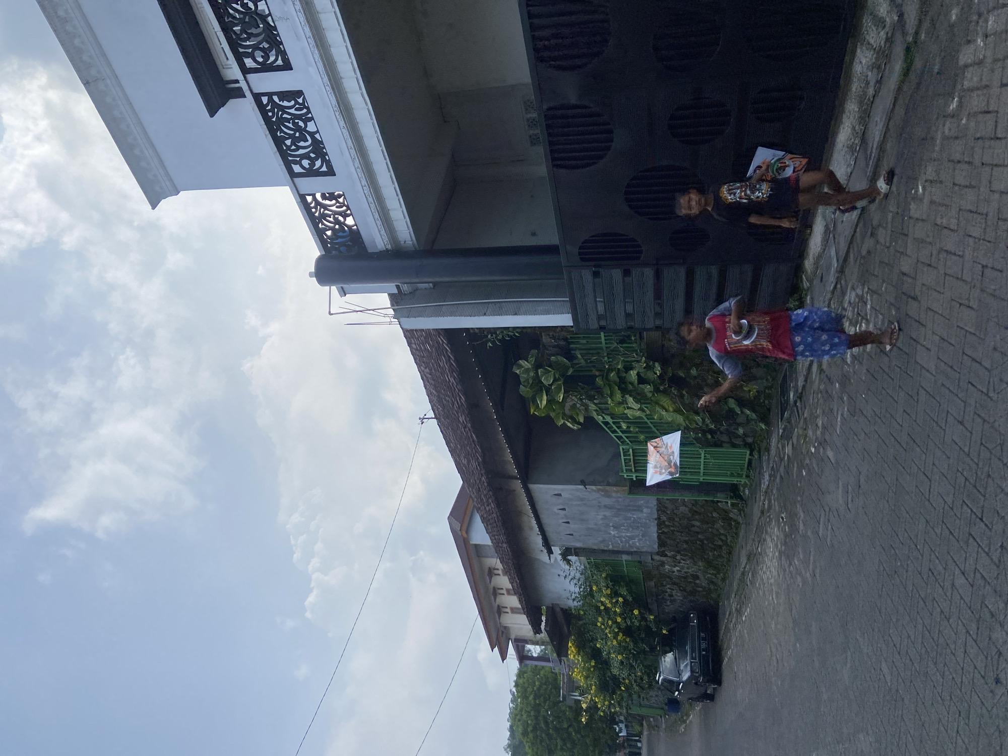 The image captures a street scene in Sukoreno, Indonesia, depicting two boys walking along a paved sidewalk. One boy wears a red and grey shirt with blue shorts, while the other wears a black shirt and shorts. They are carrying what appears to be posters or flyers. The background includes residential buildings with varying architectural styles, some featuring decorative elements. Green vegetation is visible along the side of one of the buildings. A black car is parked further down the street. The sky is overcast with light clouds.