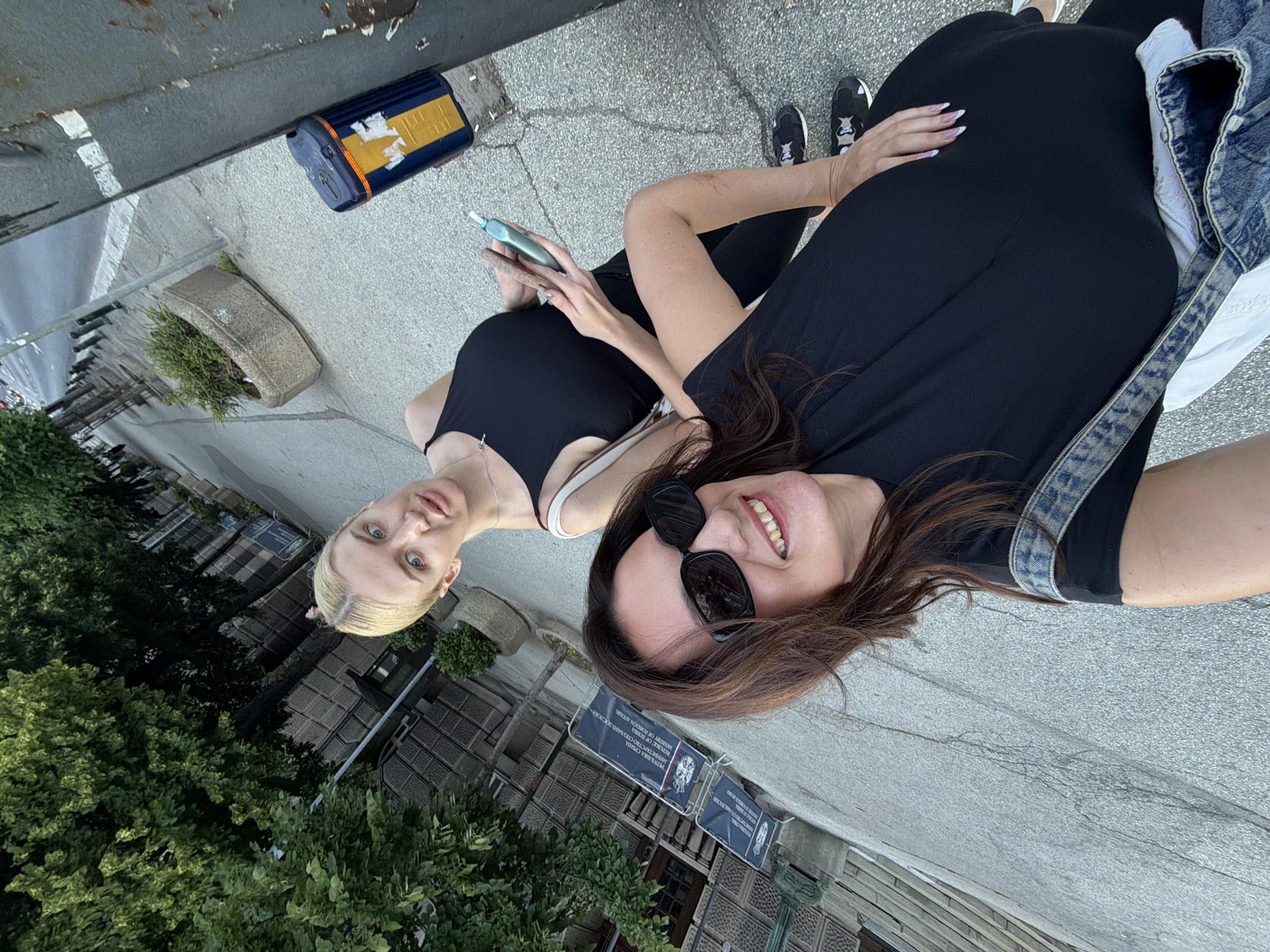 Two women are taking a selfie outdoors. One woman has blonde hair, a black tank top, and is holding a smartphone. The other woman has brown hair, is wearing sunglasses, a black dress, and a denim jacket. They are standing on a gray paved surface. In the background are trees, buildings, and signs with Cyrillic text, identifying the location as Belgrade, Serbia.