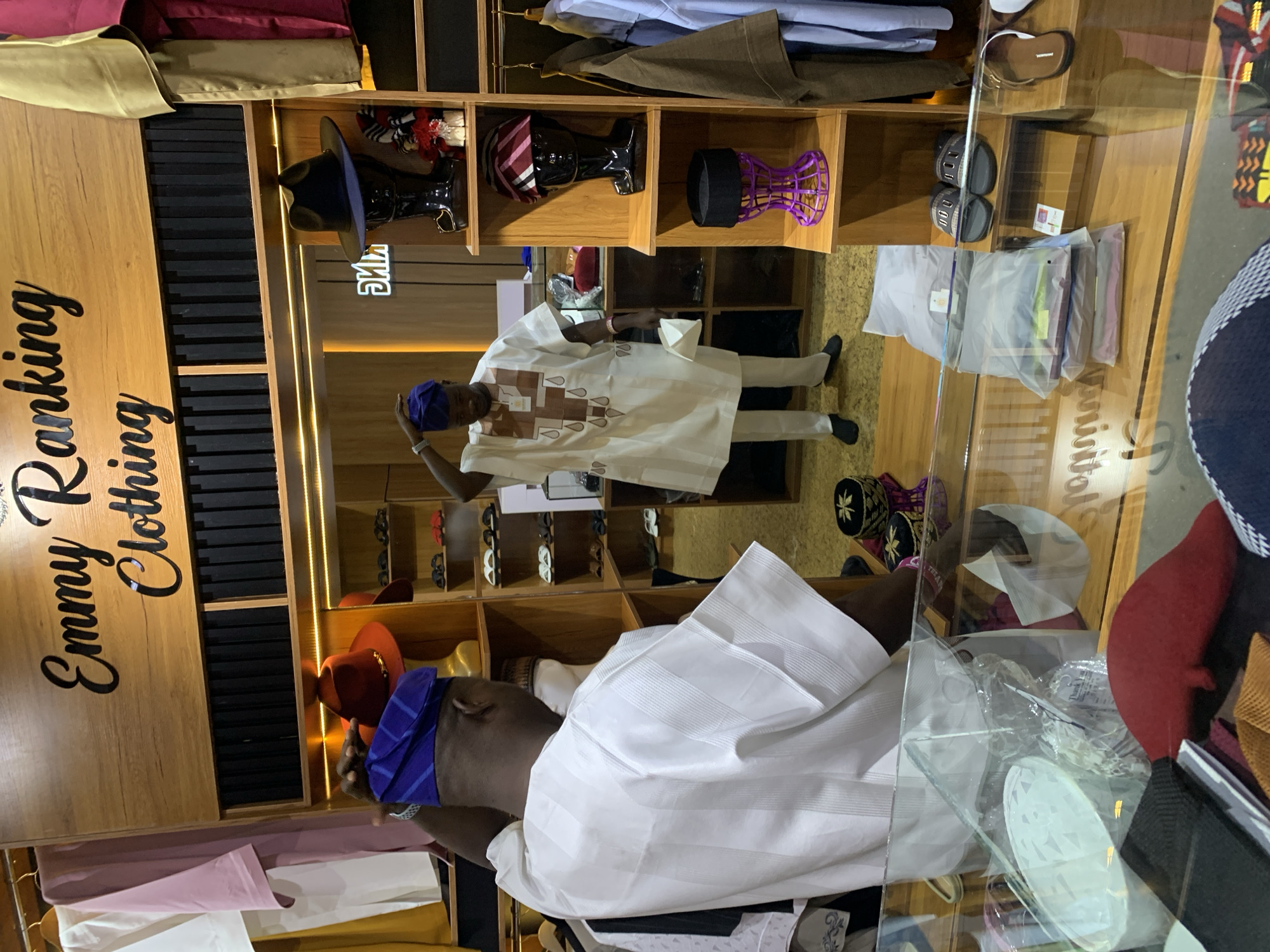 This image depicts the interior of a clothing store, identified as "Emmy Ranking Clothing" in Gura Top, Nigeria. A man, dressed in traditional white attire and a blue cap, is visible from the back, adjusting his headwear. He is standing in front of a mirror, which reflects another man in similar clothing. The store features wooden shelving units displaying various items, including hats on mannequins, slippers, and folded clothing packages. The background shows racks of clothing, and the overall environment suggests a retail setting with a focus on traditional African garments.