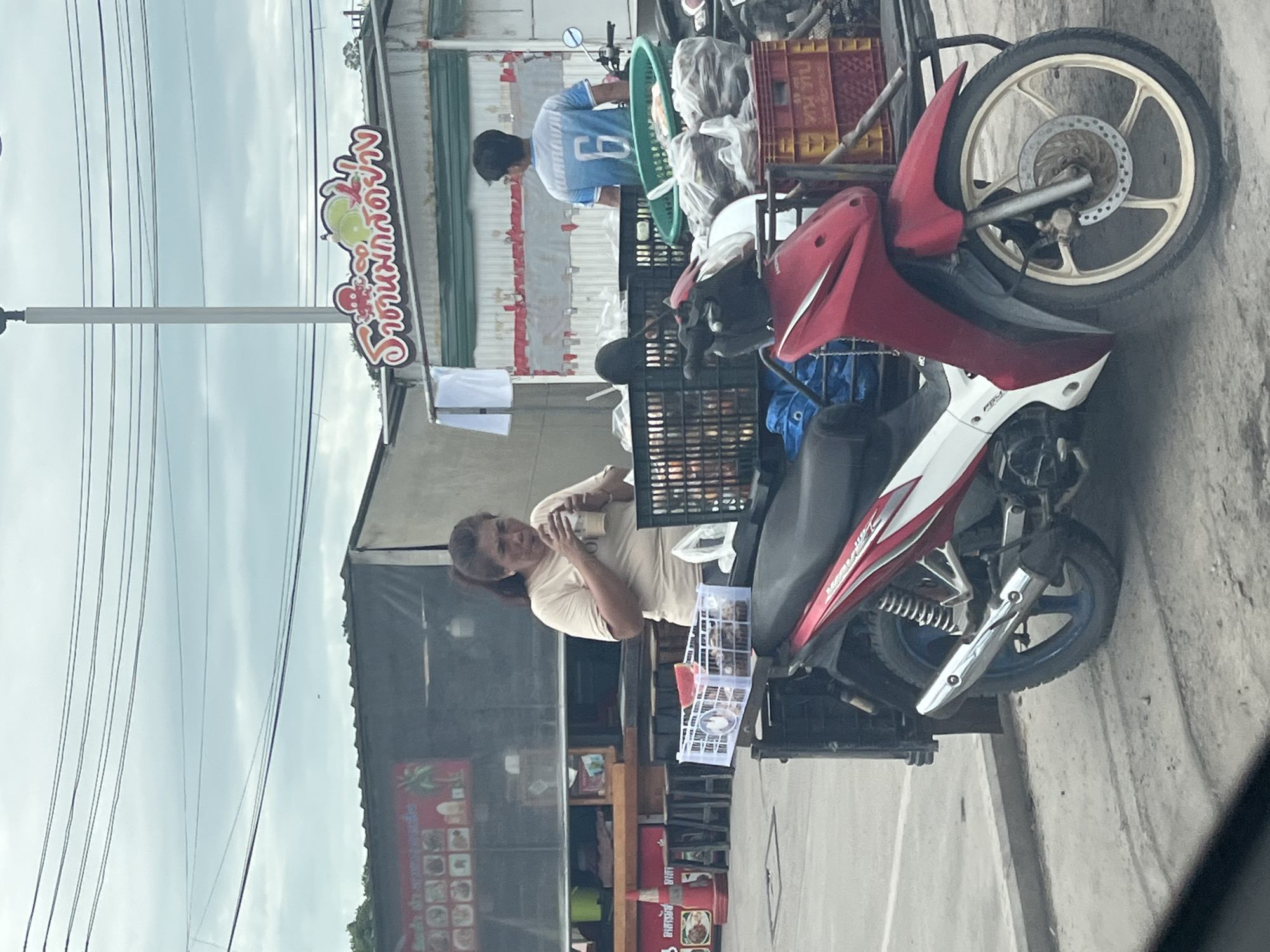 A woman in a beige t-shirt is drinking from a clear plastic cup with a straw. She is standing next to a red and white motorcycle that is loaded with goods in crates and plastic bags. Behind her, a man in a blue t-shirt with the number 6 on the back is standing. The scene appears to be a street or roadside stall in Ban Bang Rak, Thailand, indicated by the Thai script on the signage. The sky is overcast.