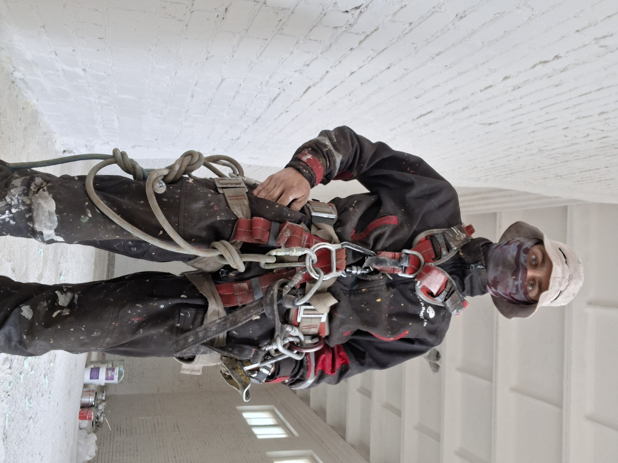 The image shows a construction worker wearing a black jacket and pants, a light-colored hat, and a face covering, standing in a building with a white brick ceiling. He is wearing a safety harness with ropes and carabiners attached. There are paint cans visible near the bottom left of the image. The location is Unknown, Unknown.