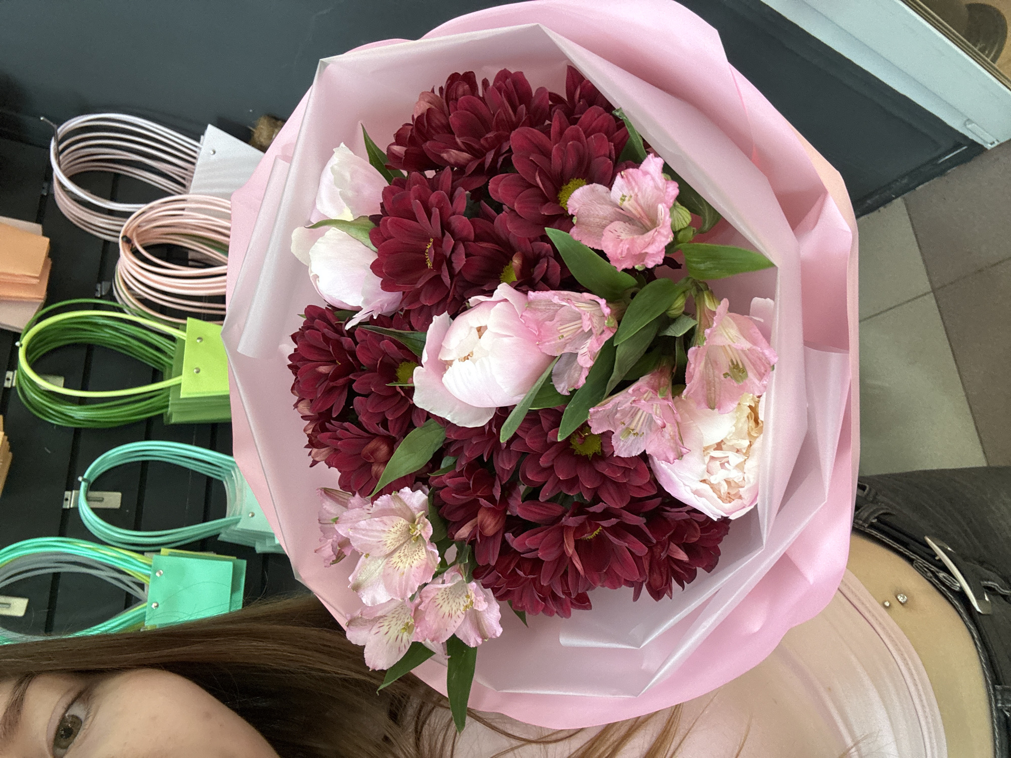 In Chaikovsky, Russia, a close-up, eye-level shot features a young woman holding a vibrant bouquet of flowers. The bouquet, predominantly pink and deep red, is wrapped in light pink paper. Among the blooms are pink alstroemerias and dark red chrysanthemums. The woman's face is partially visible on the left, her brown hair framing her face. She is wearing a pink top and dark pants, with a belly button piercing visible. In the blurred background, rows of colorful, looped gift bag handles in shades of green and pink are attached to a dark wall. The lighting is bright, illuminating the flowers and the woman's upper body.