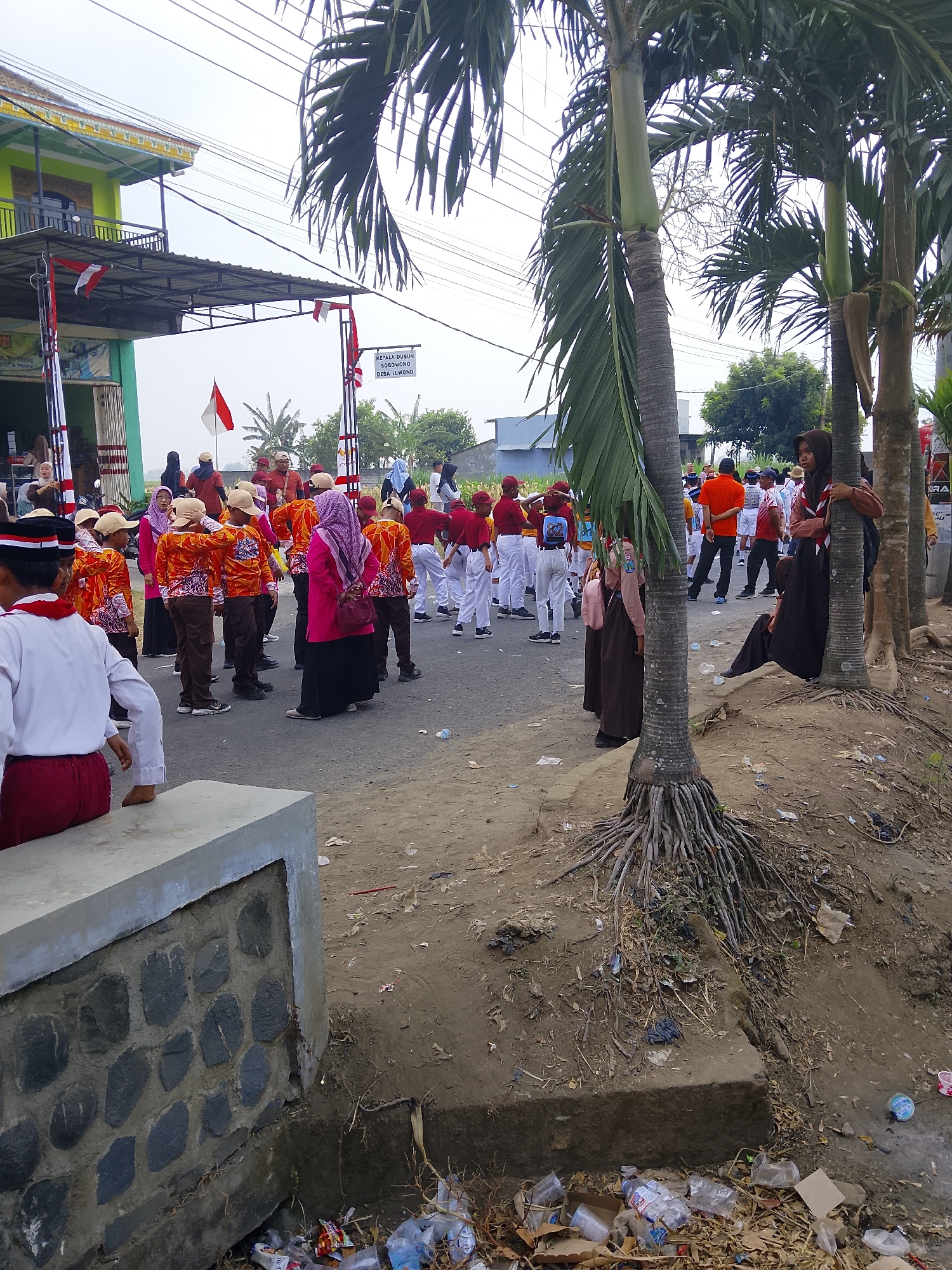 The image depicts a scene in Bangsri, Indonesia, likely during a local parade or event, as evidenced by the presence of children in uniform, some carrying flags. In the foreground, a person in a white and red uniform is seated on a concrete structure, facing away from the camera. A group of children in matching orange shirts with patterned sleeves and brown pants, some wearing caps, are gathered in the middle ground. Several adults are also present, some in orange shirts and others in traditional Indonesian attire. Palm trees with exposed root systems line the right side of the frame, casting shadows on the ground. The environment appears to be a street or a dusty area adjacent to a building, with some debris and plastic bottles scattered on the ground. In the background, there are more buildings, trees, and power lines. The overall atmosphere suggests a community gathering or celebration.