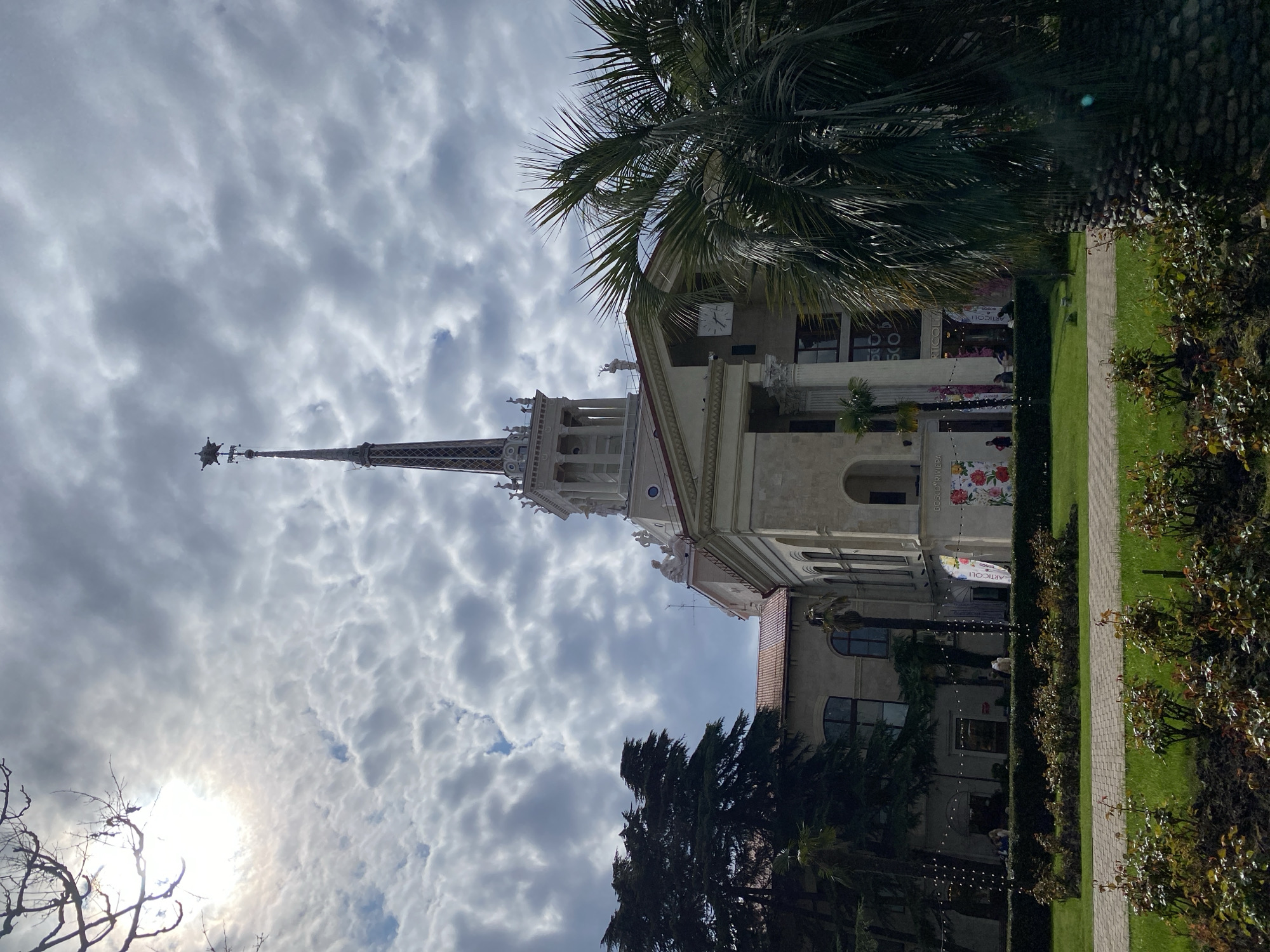 The image depicts a tall, ornate building in Sochi, Russia, likely a landmark structure. The building features a prominent spire and classical architectural elements such as columns and statues. A clock is visible on the building's façade. The building is partially obscured by a palm tree on the right side of the image.
The sky is overcast with textured clouds. The sun's position is indicated by a bright area in the lower left corner.
The foreground includes greenery such as shrubs and grass. A paved walkway extends along the building's side. Several people are present, likely tourists or locals visiting the area. The atmosphere suggests a pleasant, possibly historical or cultural site.