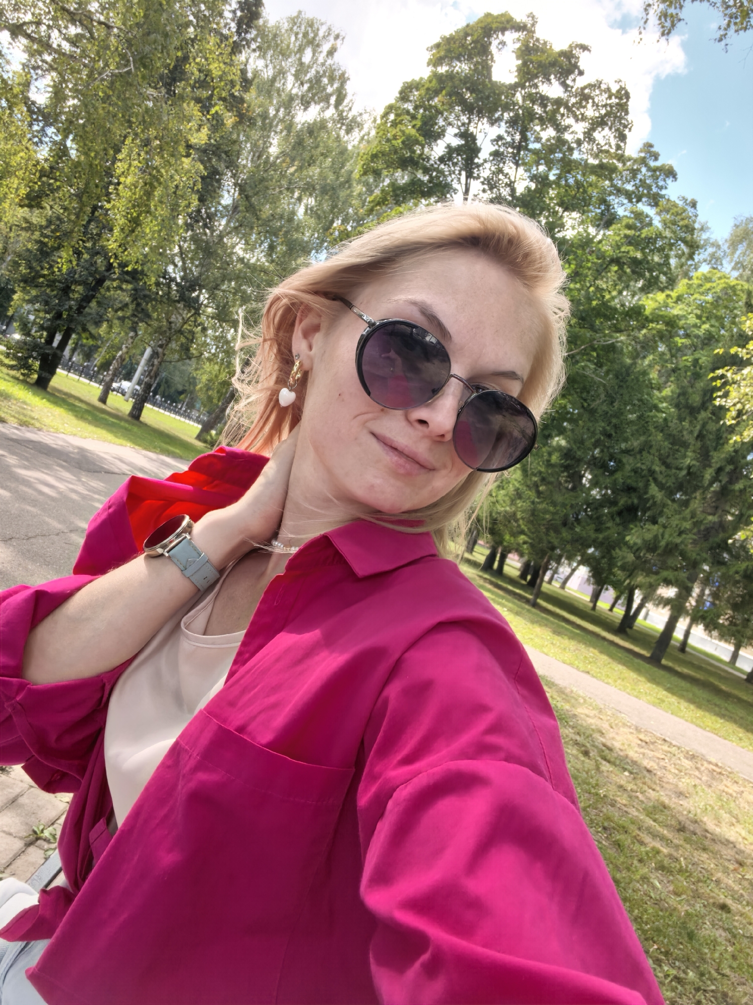 A woman is taking a selfie outdoors in a park. She has blonde hair and is wearing large, round sunglasses, a bright pink button-up shirt, and a cream-colored tank top underneath. A light blue watch is on her left wrist. She is wearing a necklace with small white pearls and gold earrings, one of which is a heart-shaped pearl. The background shows trees, a pathway, and a grassy area. The sky is blue with some clouds. The image appears to have been taken on a sunny day. The location is identified as the city of Sterlitamak, Russia.