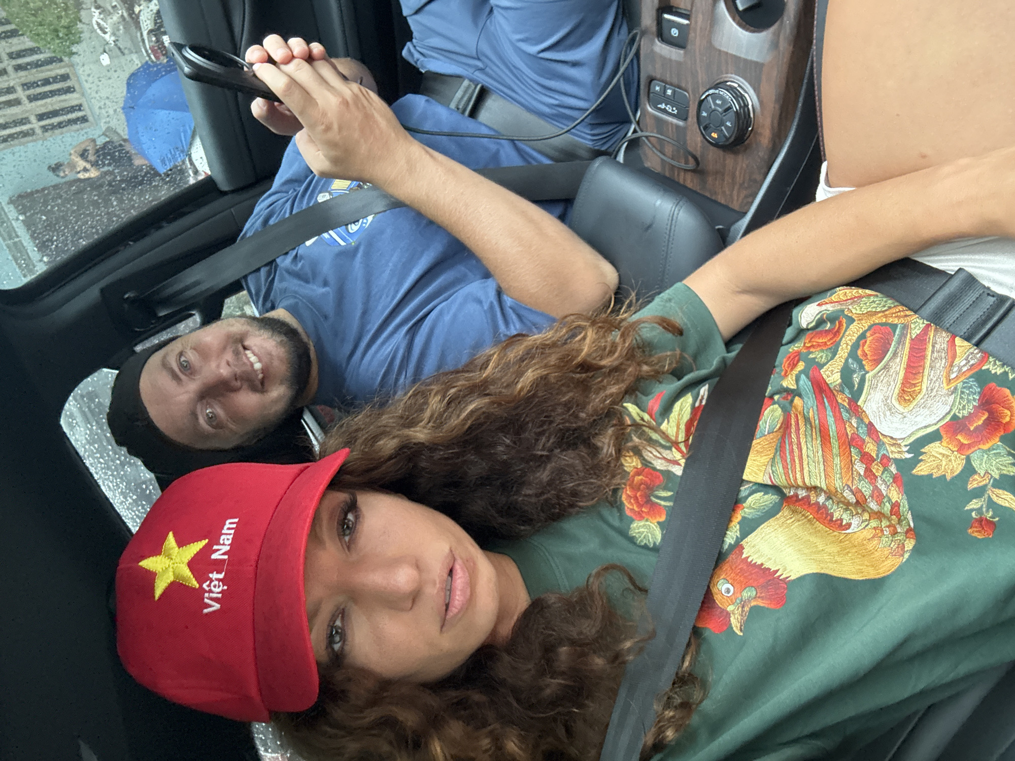 A man and a woman are sitting in a vehicle. The woman is wearing a red baseball cap with a yellow star and the words "Việt Nam" embroidered on it, and an olive green shirt with a design featuring a rooster and flowers. The man is wearing a blue t-shirt and holding a black smartphone. Both are wearing seatbelts. Rain can be seen on the window of the vehicle. The interior of the vehicle includes wood trim and various controls. Based on the Vietnamese writing on the hat, it is likely that the image was taken in Vietnam, but there is no evidence from the image to suggest the exact location of Nghia Lo Ward.