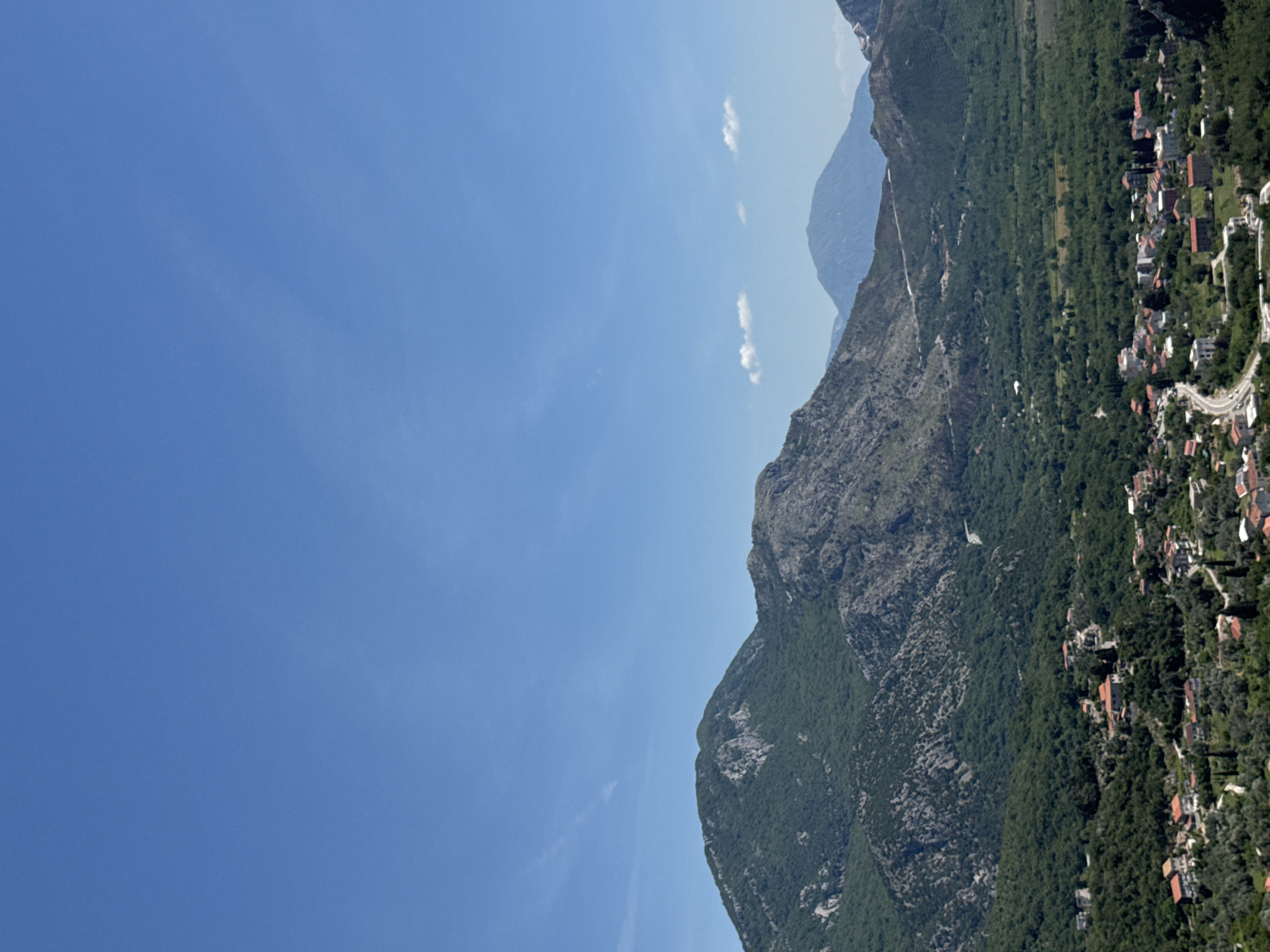 The image depicts a view in Petrovac na Moru, Montenegro. A mountain range, partly covered in lush vegetation, dominates the right side of the frame. A cluster of buildings, presumably a residential area, is visible nestled amongst the trees at the mountain base, with roads winding through it. The sky, a gradient of blue, occupies the majority of the left side, with a few wisps of clouds scattered about.