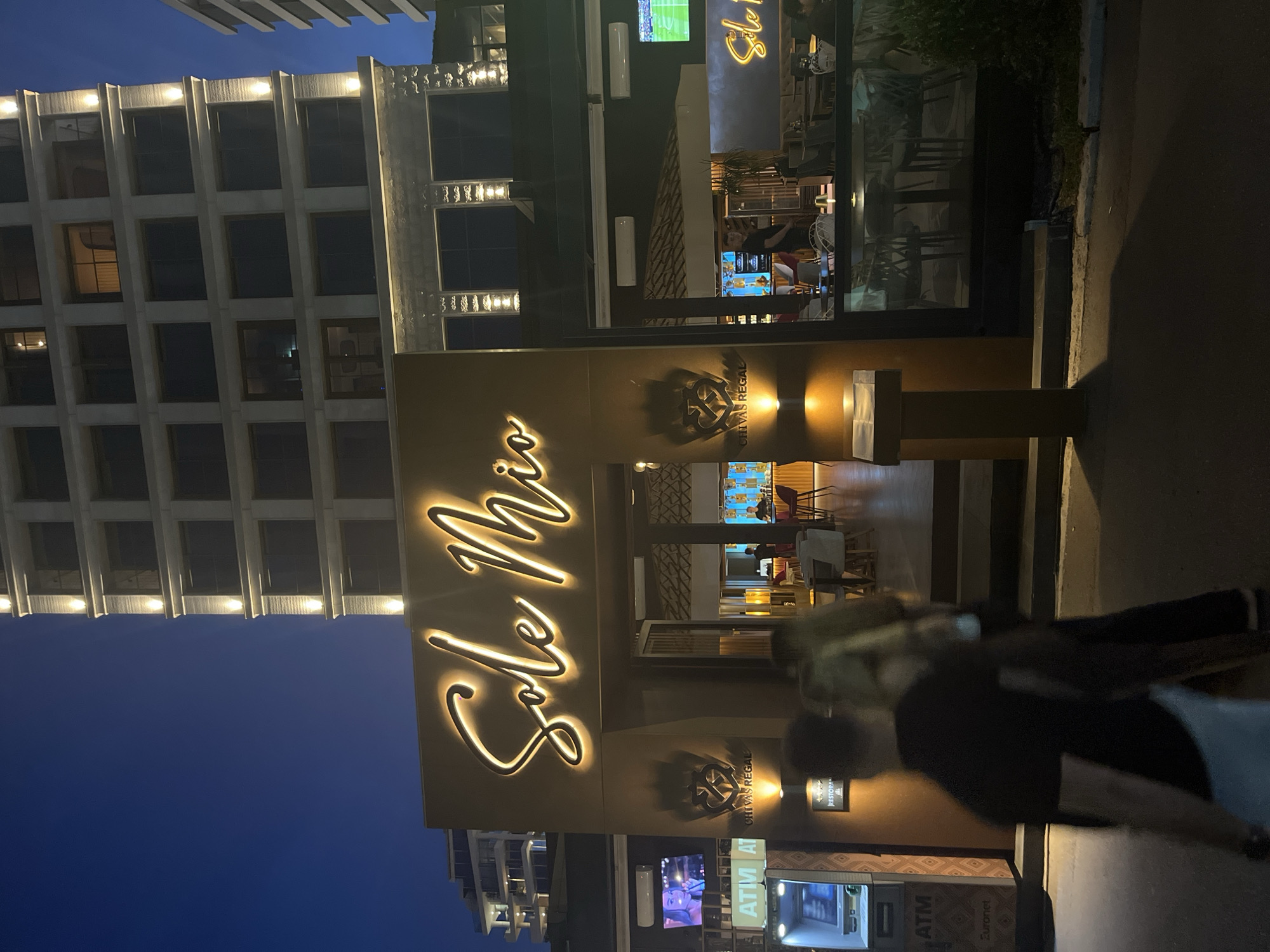 The image depicts a nighttime scene in an urban environment, likely outside a restaurant or bar. The focal point is a building with a large illuminated sign that reads "Sole Mio" in a cursive font. Below the sign, the entrance to a business is visible, with interior lighting illuminating tables and chairs. The building itself appears to be modern, with multiple floors and numerous windows. To the left of the entrance, there is an ATM machine and a television screen displaying an image of a woman. Two ATMs are present. In the foreground, the blurry figures of two people walking are visible, suggesting a public space. The sky is dark, indicating it is nighttime. The overall scene suggests a lively, commercial area.