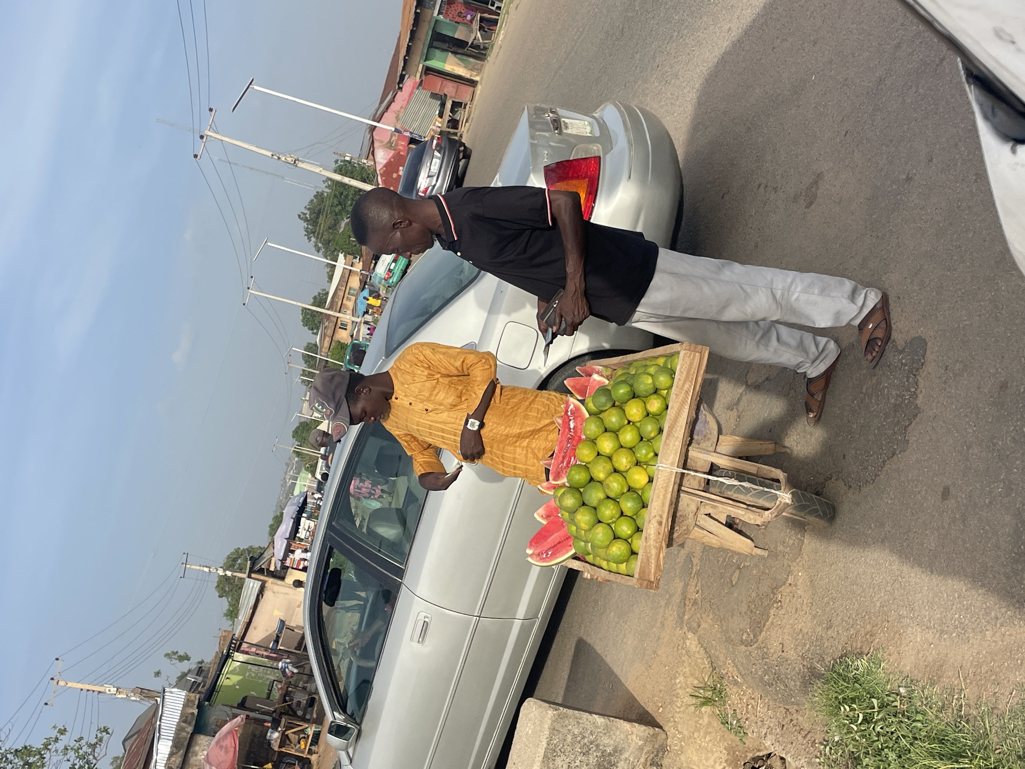 The image shows two men beside a silver car on a street in Jos, Nigeria. One man wears a black shirt and light-colored pants, looking at his mobile device. The other man, in a mustard-colored long-sleeved shirt and cap, stands beside a wooden cart filled with green oranges and cut watermelons. The cart appears to be handmade with a single wheel. The scene is roadside, with shops and buildings in the background. Overhead power lines are visible against a bright sky. The road shows signs of wear and tear.