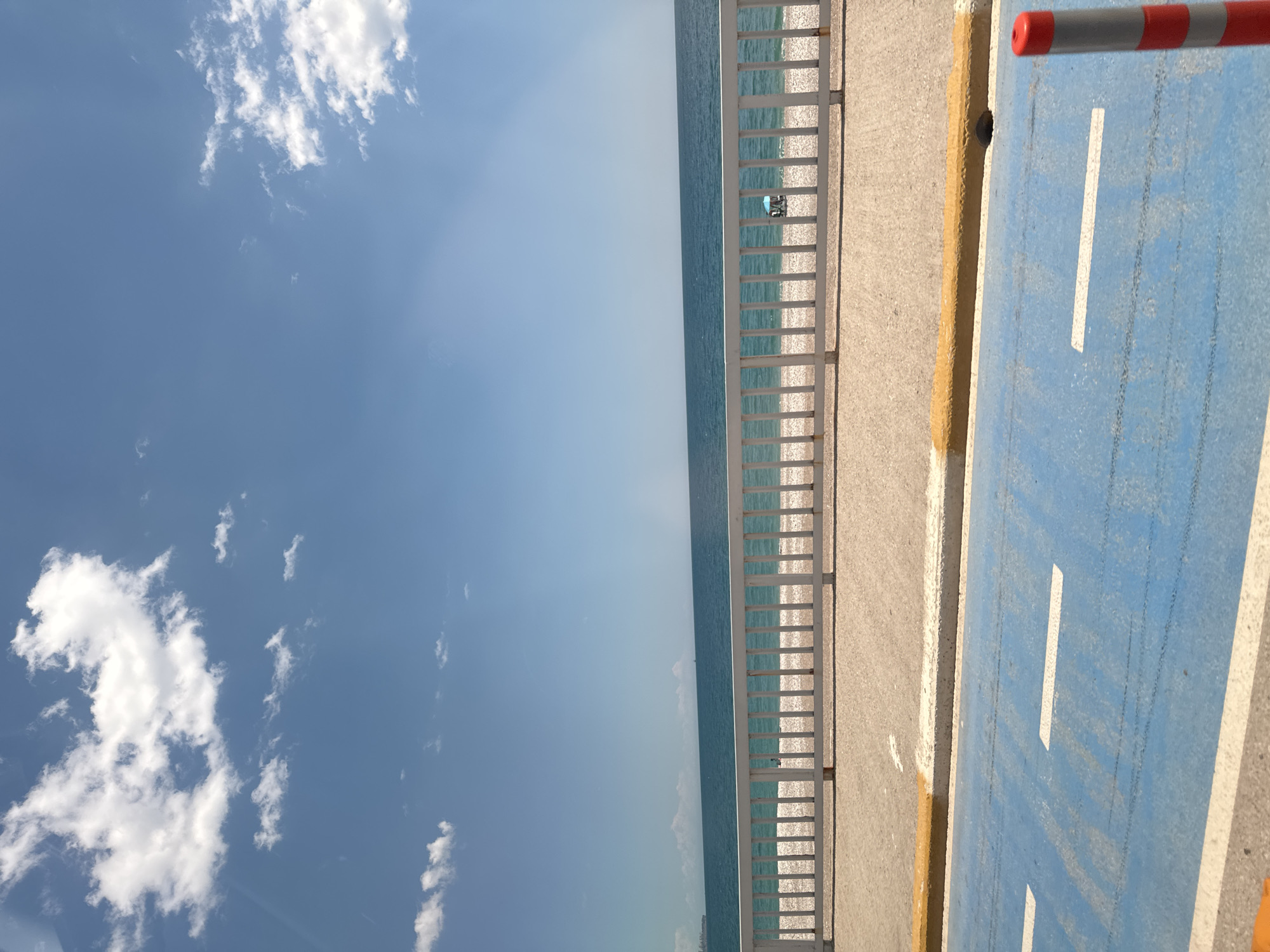 The image depicts a sunny day at Konyaaltı Beach in Antalya, Turkey. A blue bike lane, marked with white stripes, runs parallel to a concrete walkway bordered by a white railing. Beyond the railing is the sea, reflecting the sunlight. The sky is blue with scattered white clouds. A beach umbrella is visible in the distance. A barrier pole, painted red and white, is partially visible on the right edge of the frame.