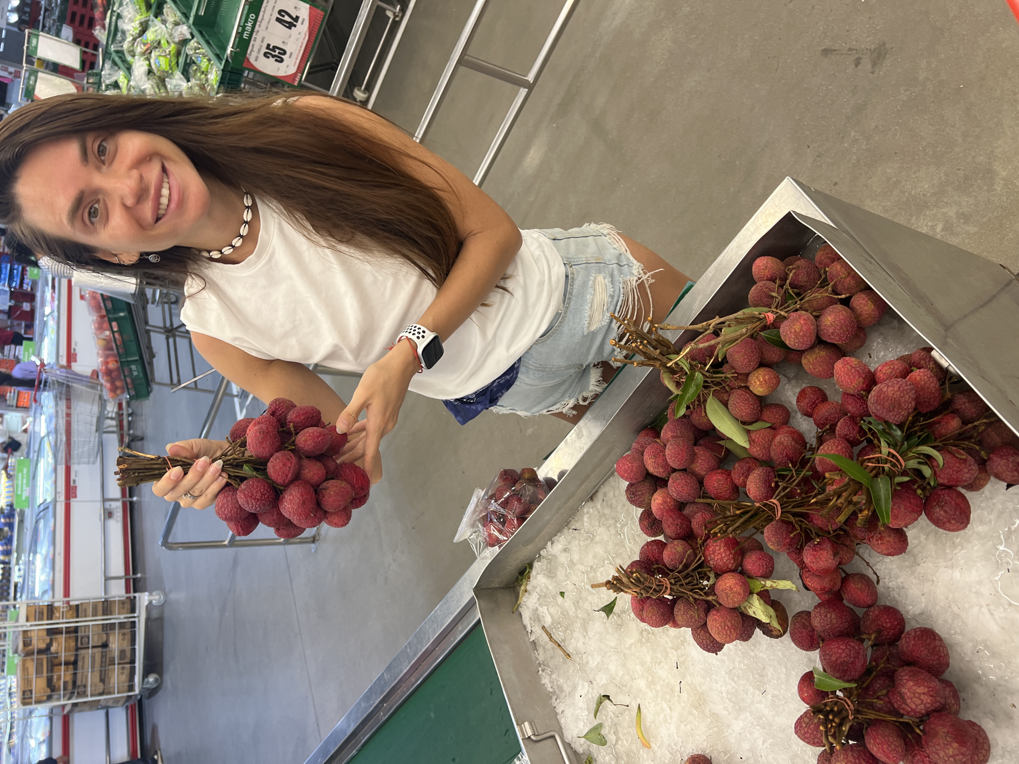 A woman stands in a supermarket in Baan Bo Phut, Thailand, holding a bunch of lychees. She is wearing a white sleeveless top, ripped denim shorts, and a seashell necklace. A smartwatch is visible on her left wrist. She is smiling and looking at the camera. In front of her, a metal display bin is filled with ice and lychees. Behind her, shelves stocked with products and shopping carts are visible. The overall environment suggests a typical supermarket setting.