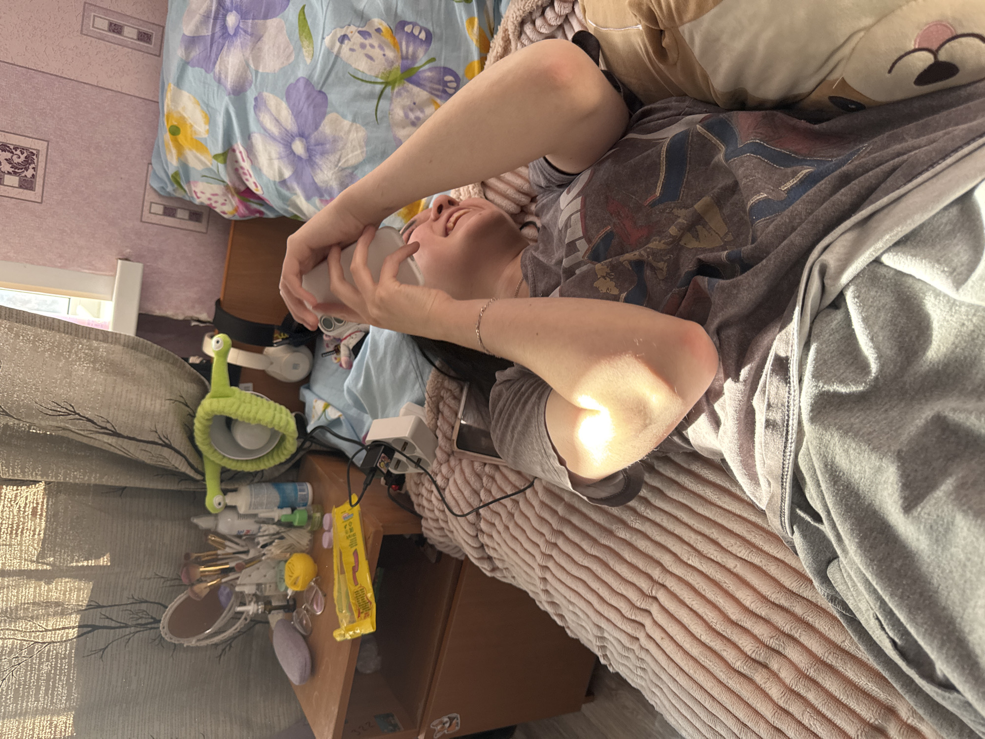 The image depicts a relaxed indoor scene in Perm, Russia, featuring a person lying horizontally on a bed, illuminated by strong sunlight. The individual, likely female, is wearing a grey t-shirt with a graphic design and grey sweatpants. She holds a white phone or small device with both hands, looking upwards with a broad smile, suggesting happiness or amusement. A silver bracelet is visible on her right wrist.
The bed is adorned with a light blue duvet patterned with purple and yellow flowers and butterflies, and a soft, ribbed pinkish-beige blanket. A plush toy, resembling a dog or bear, is used as a pillow.
Adjacent to the bed, a wooden nightstand is cluttered with personal items. These include a distinctive green flexible desk lamp shaped like a cartoon snail, various cosmetic bottles and tubes, makeup brushes in a holder, a small mirror, eyeglasses, a purple puff, and a yellow packet (likely snacks). A white power strip with USB ports and cables, along with white headphones featuring green accents, are also present. The numbers "332" are faintly visible on the side of the nightstand.
Strong sunlight streams through a window, casting distinct shadows on the light-colored curtains, which are patterned with dark branches. The wall features light pink wallpaper with subtle rectangular designs. The bright, natural light suggests it is either morning or late afternoon. The overall atmosphere is casual and comfortable.