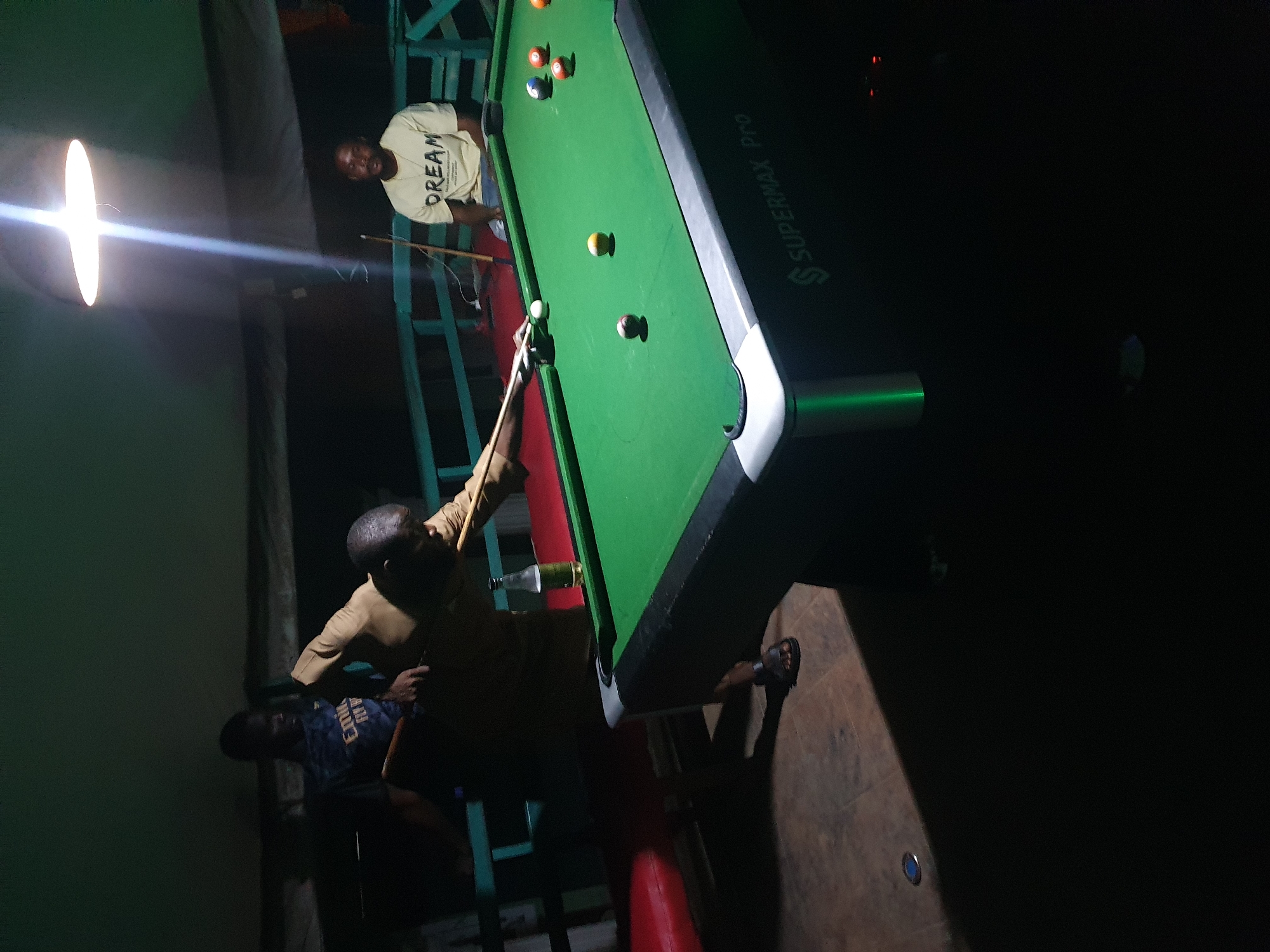 The image depicts an indoor scene at night, likely in Lokoja, Nigeria, where a group of men are playing pool. The primary focus is a pool table with green felt, illuminated by a bright overhead light. Several pool balls are scattered across the table, including a cue ball, a yellow ball, and a red and white striped ball.
Three men are visible. One man, wearing a light-colored t-shirt with the word "DREAM" printed on it, is standing near the table, holding a pool cue. Another man, wearing a dark t-shirt with yellow lettering that appears to say "EARLY," is seated on a bench or chair. A third man, wearing a light brown shirt, is in the process of taking a shot, aiming his cue at the white ball.
The environment appears to be casual, with what looks like wooden flooring and some green metal structures that might be part of the building's framework or seating. A couple of glass bottles, one with a green liquid, are visible on the side of the pool table, suggesting a relaxed social gathering. The lighting is somewhat dim, with the bright overhead light creating strong contrasts and shadows. The overall impression is that of a friendly game of pool taking place in the evening.