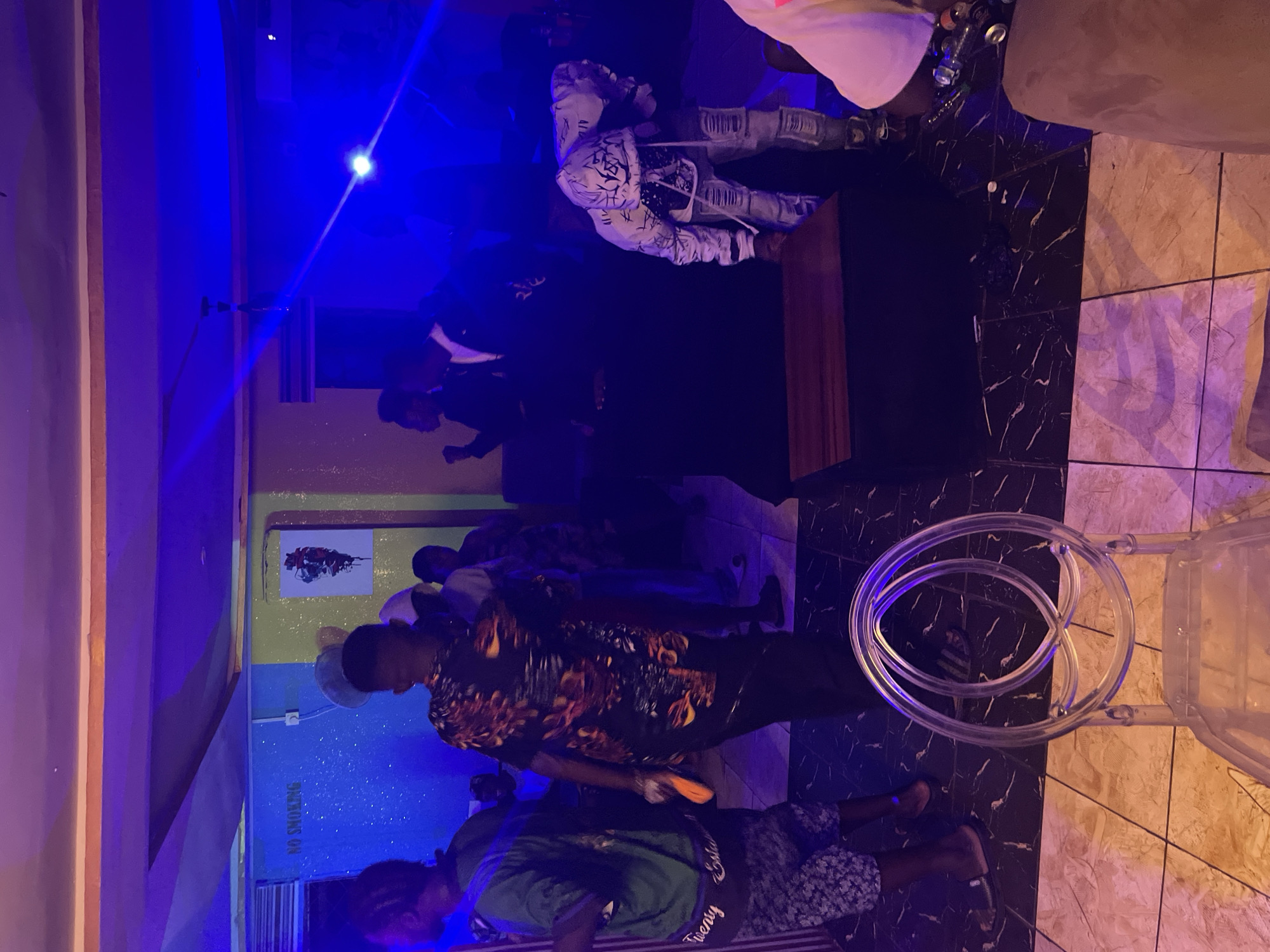 The image shows an indoor scene in what appears to be a bar or club, possibly in Durimi, Nigeria. Several people are present, with one person in a black and orange patterned shirt standing prominently. A person in a black suit is also visible. There's a "NO SMOKING" sign on the wall, which is painted in shades of blue and yellow. A clear chair is visible in the foreground. The lighting is blue, creating a nightclub atmosphere. The floor is tiled in a mix of dark and light marble patterns.
