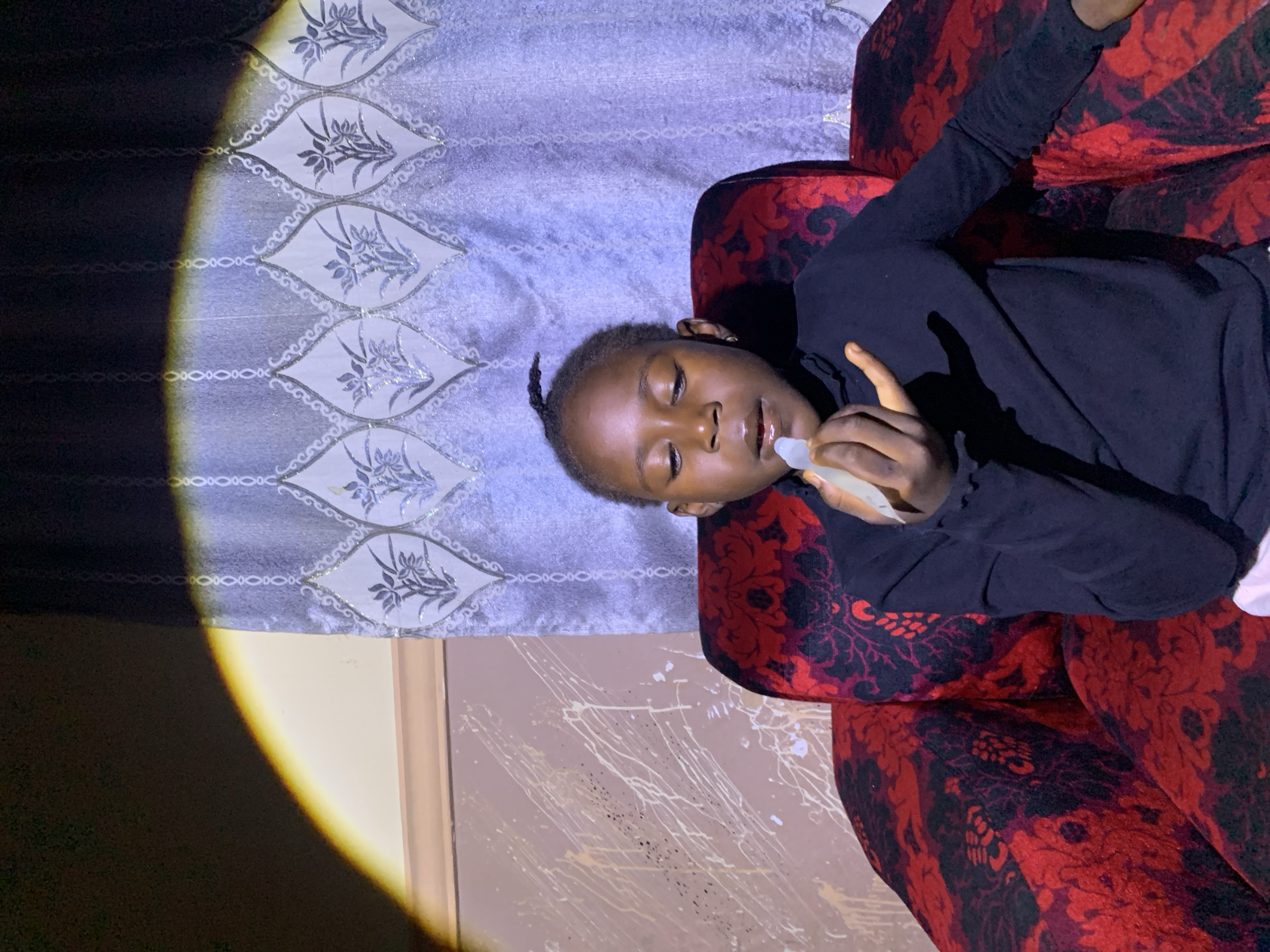 A young Black girl with dark skin, braided hair pulled back into a ponytail, and wearing a black turtleneck sits in a large, ornate red armchair. The armchair has a black floral pattern. She is holding a small, clear tube with a white applicator to her lips, appearing to apply lip balm. Her eyes are closed, and her head is tilted slightly downwards. The background consists of a wall with a textured, splattered paint design and a light purple curtain with a silver floral pattern. A bright, circular spotlight illuminates the left side of the scene, casting a shadow on the girl's arm and the chair. The scene appears to be indoors, likely in a residential setting. There is no information that allows for the precise location in Russo, Nigeria to be determined.