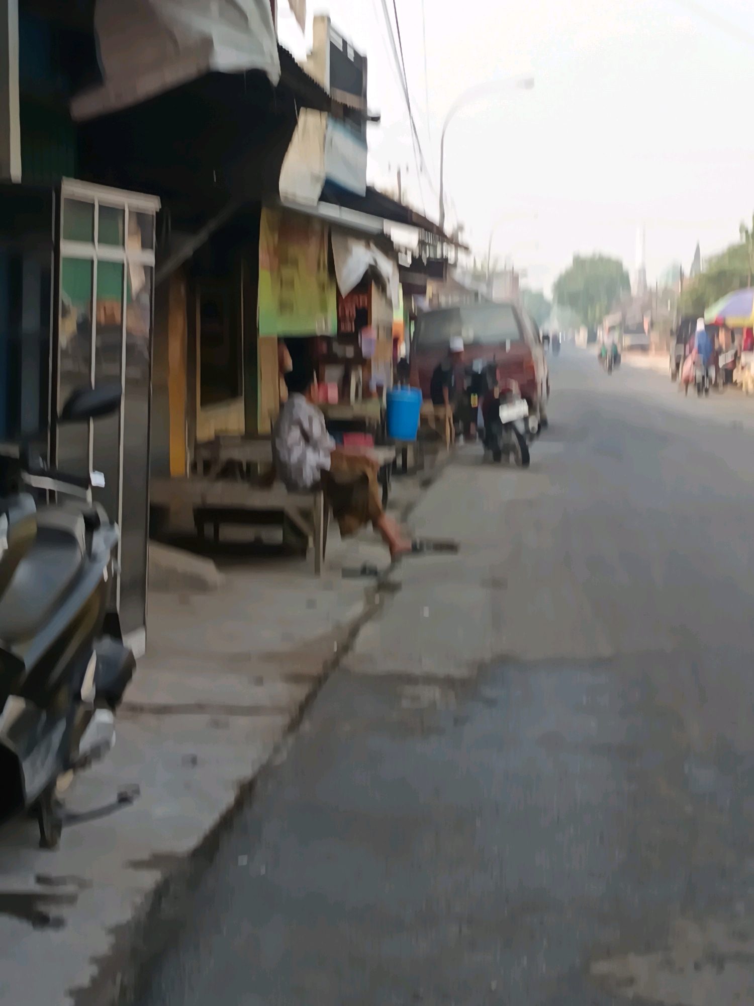 The image depicts a street scene in Blega, Indonesia. On the left side of the frame, there is a building with a white-framed window and a motorcycle parked in front of it. A person is seated on a wooden bench outside a shop on the left side of the street. To the right of the person, a red van is parked on the side of the road. Further down the street, other vehicles, including motorcycles and a cart with an umbrella, are visible. The road appears to be paved and wet in some areas. The environment suggests a typical urban or semi-urban setting in Indonesia, possibly a market or roadside stall area.