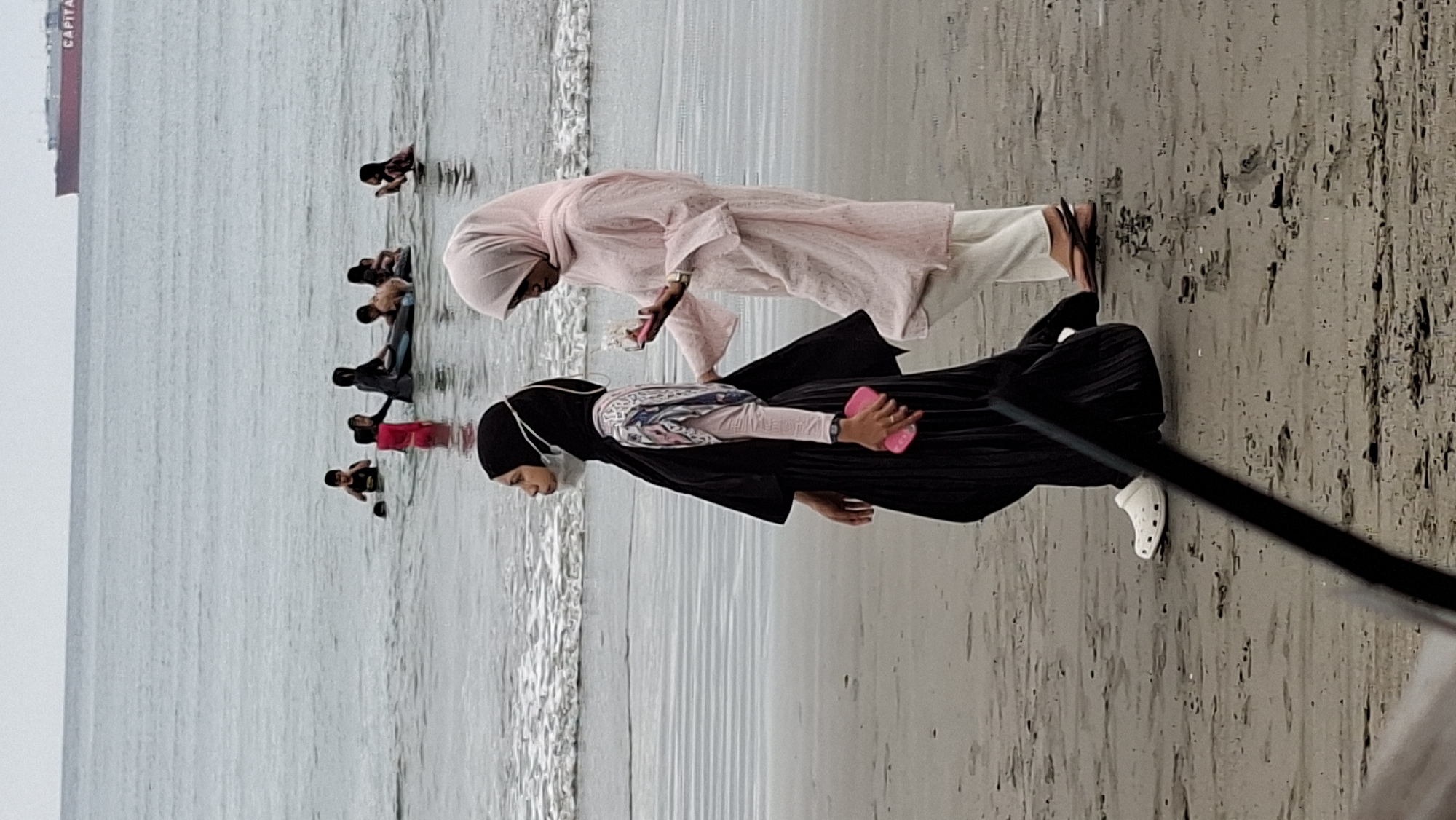The image captures a cloudy day at a beach, likely in Lhokseumawe, Indonesia. In the foreground, two women are walking on the sand. The woman on the left wears a black hijab, a light-colored patterned shirt, and a black pleated skirt, with a face mask over her nose and mouth. She is also wearing white clogs. The woman on the right wears a pale pink hijab and a long, loose-fitting pink tunic over white pants, with brown sandals. She is holding a pink mobile phone in her right hand.
In the background, several children are playing in the ocean. Some are wading, while others are in a large inflatable ring. Further out in the sea, a large red and white cargo ship with "CAPITAL" visible on its side is anchored. The water is choppy with visible waves breaking on the shore. The sand is damp and shows some footprints. The overall scene depicts a casual outing at the beach.