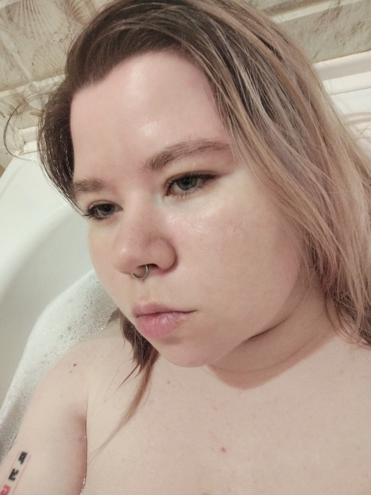 A fair-skinned person with light brown hair is partially submerged in a bathtub filled with soapy water. The person is looking down and to the left. They have a silver hoop in their septum. A tattoo is visible on their left arm. The location is a bathroom, implied by the bathtub.