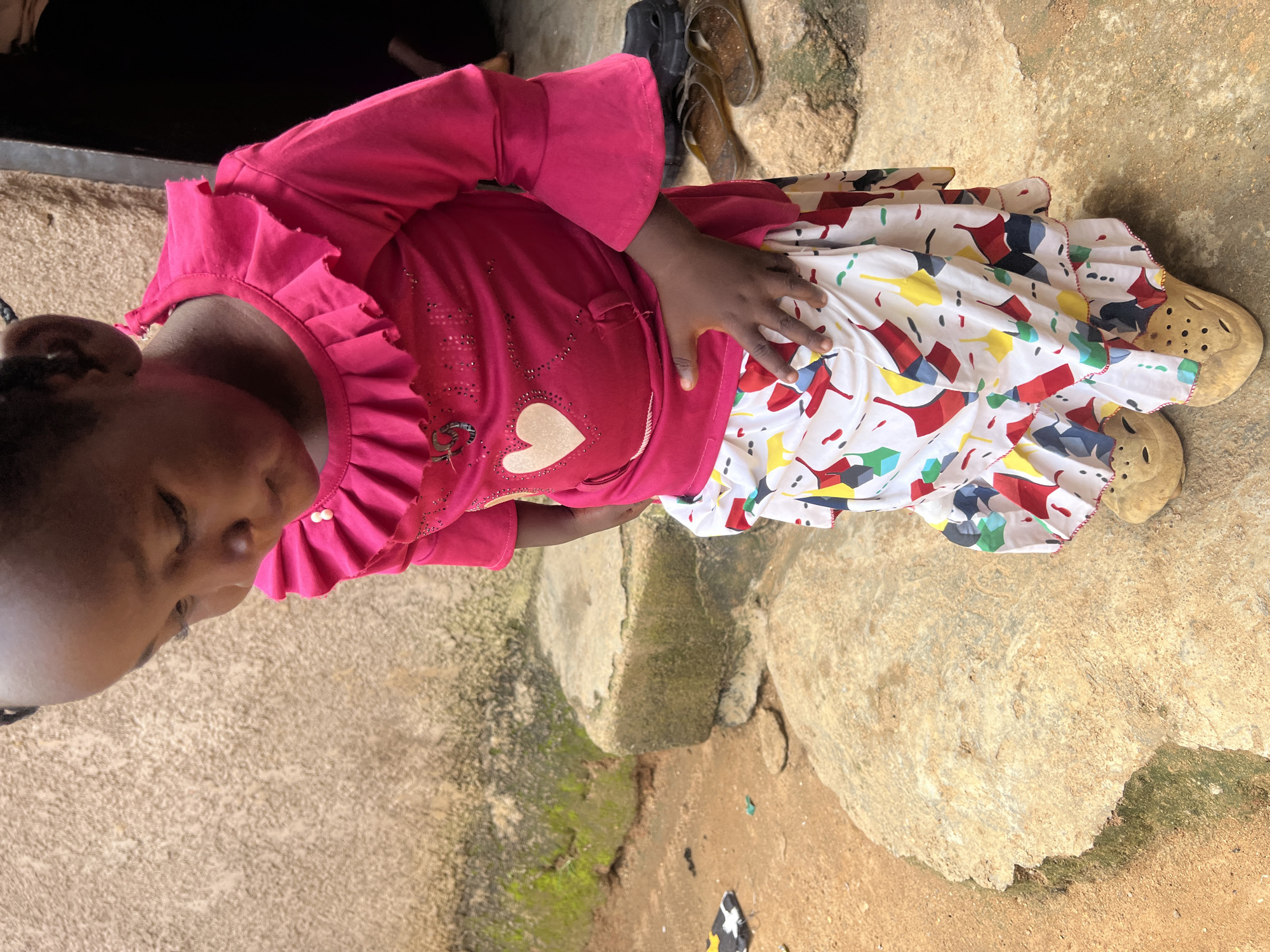 A young child, seen from the chest down, is lying on a rough, textured, tan-colored surface. The child is wearing a vibrant pink long-sleeved shirt with ruffled details around the neckline and a skirt adorned with a colorful, abstract pattern of geometric shapes in red, yellow, blue, and black on a white background. The child's right hand is resting on their stomach, fingers slightly splayed. On their feet, they wear light-colored sandals with a perforated design.
The background consists of more of the same textured ground, with patches of mossy green suggesting dampness or uneven surfaces. In the upper portion of the image, a dark, blurry shape, possibly furniture or clothing, is visible. The lighting suggests it could be daytime. The scene appears to be outdoors, possibly in a dusty or unpaved area given the ground texture. The overall impression is casual and candid. There is no visible text in the image.