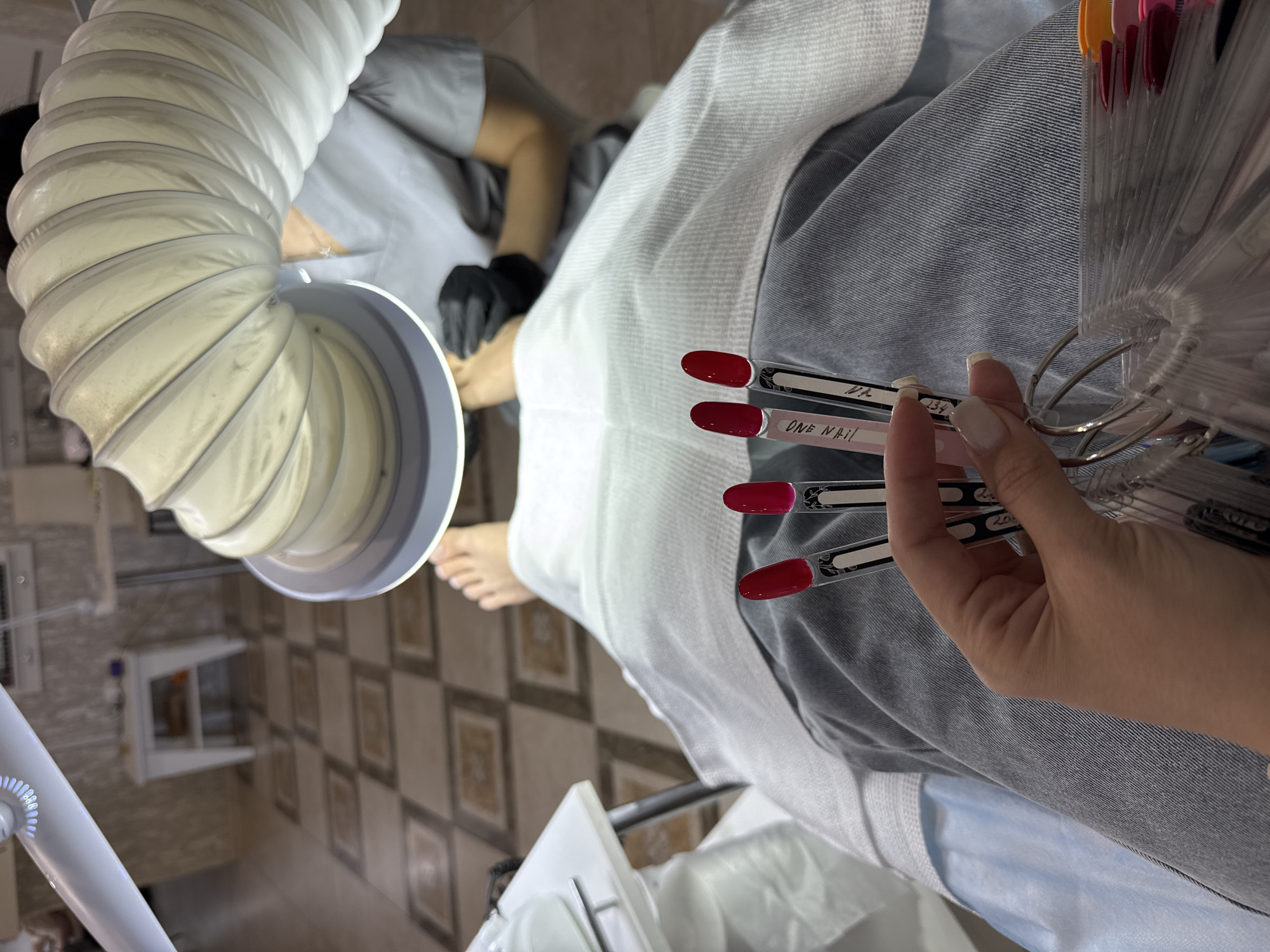 The image shows a close-up view of a person holding a set of nail polish sample sticks, with several red and pink shades visible. The sticks are on a ring, suggesting a larger collection. In the background, a foot is being worked on by a person wearing black gloves and a gray uniform, likely a nail technician in a salon. There's a large, flexible hose connected to a light fixture positioned above the foot. The environment appears to be a nail salon, with tiled flooring and white furniture in the background. The image's perspective suggests the viewer is either the client or someone sitting opposite the nail technician. The location is identified as Minusinsk, Russia, though no specific visual cues in the image confirm this.