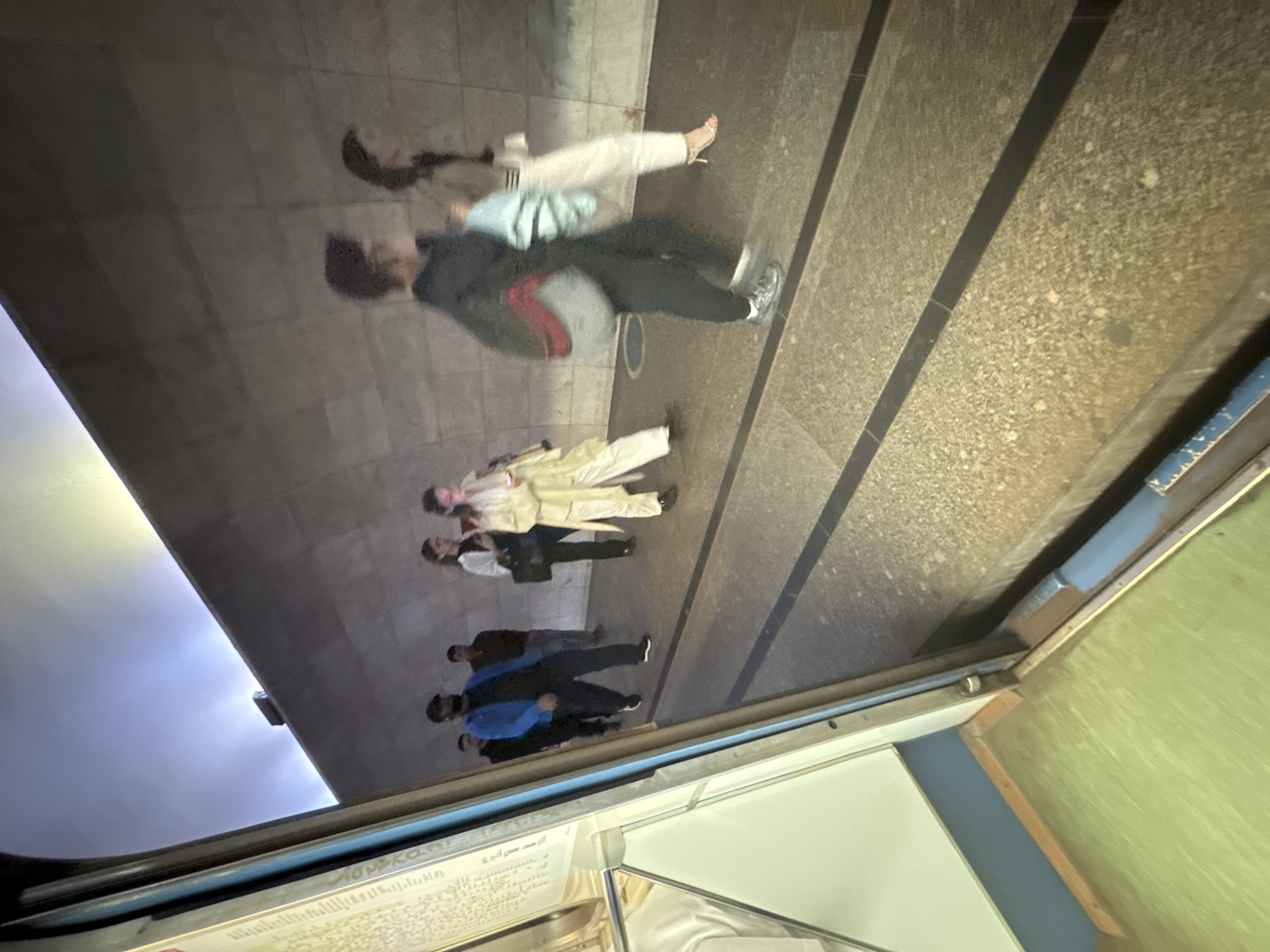 The image depicts a group of people walking along a platform, likely in a Moscow Metro station. The environment features a concrete wall with subtle texturing and a tiled floor with dark linear accents. In the foreground, part of a train car's interior is visible, with a green floor and a blue trim. The people are casually dressed, some carrying bags. The lighting suggests an indoor, possibly underground, setting. One individual in the foreground wears a blue jacket and a black cap, while others are walking in the same direction. The overall atmosphere is that of a public transit space during a journey.