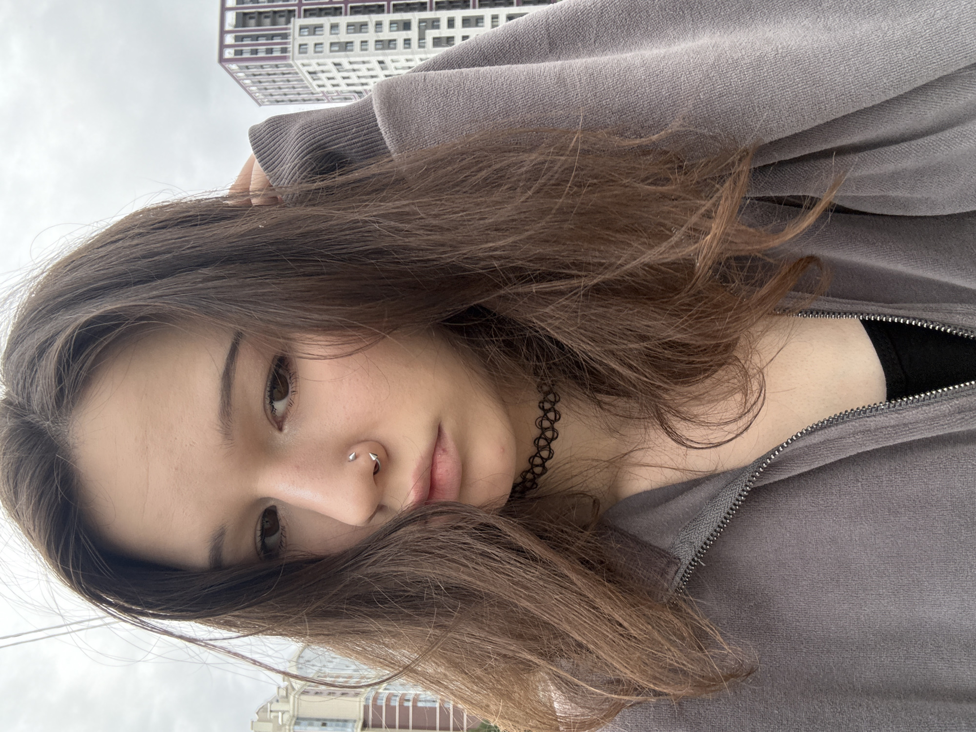 A young Caucasian woman with long, brown hair is posing for a selfie outdoors, with a slightly overcast sky and modern buildings in the background. She is wearing a gray zip-up hoodie over a black garment and a black choker necklace. She has a nose ring with two small, pointed studs. The buildings in the background appear to be residential or office buildings, with a light color scheme and purple accents. The overall scene suggests an urban or suburban setting in Kokkorevo, Russia.