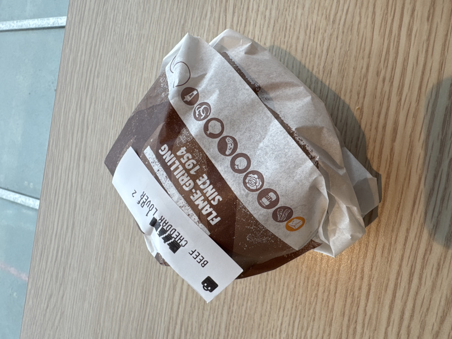The image displays a single wrapped food item resting on a light-colored, wood-grain table surface. The item is predominantly covered in dark brown paper with white text stating "FLAME-GRILLING SINCE 1954." A lighter, off-white paper strip wraps around the item, featuring circular icons depicting various food ingredients such as bacon, mayonnaise, lettuce, tomato, pickle, and onion. A white label with black text protrudes from the top, reading "BEEF CHEDDAR LOVER 2," accompanied by a shopping bag icon. The background reveals a light blue or grey tiled floor with visible white grout lines. This scene is located within a Burger King restaurant at Via Giusti, 12, 21019 Somma Lombardo VA, Italy.