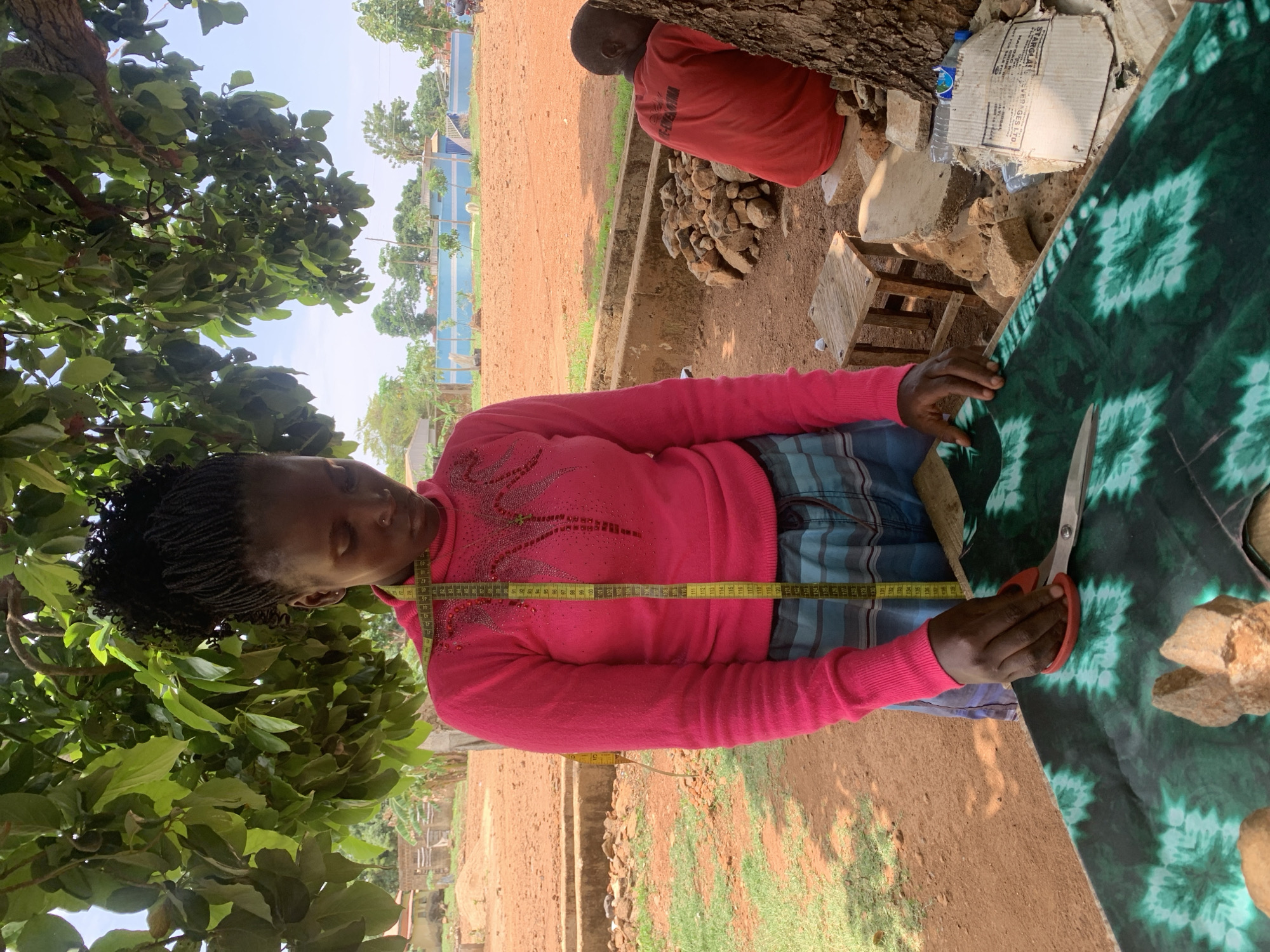 A woman with braided hair is standing outside, holding a pair of scissors in her right hand, cutting a green and blue patterned fabric. A yellow measuring tape is draped around her neck, over a pink sweater. The ground is dirt, and there is a tree to the left of the woman. To the right, partially visible, is a person sitting in a red shirt. A piece of cardboard, water bottle and some rocks are behind the seated individual. The location is the city of Maigizo, Nigeria.