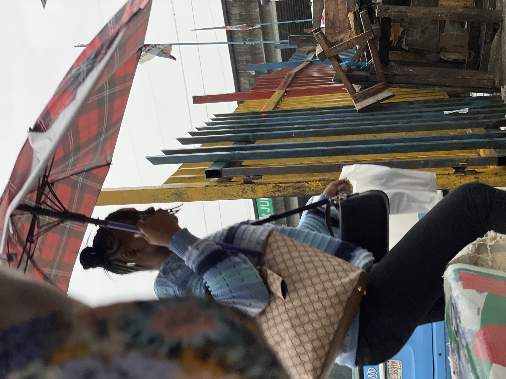 A low-angle, medium shot shows a Black woman walking under a red and black plaid umbrella in Lekki, Nigeria. The woman is wearing a light blue and white striped sweater and black pants, carrying a beige designer handbag on her left shoulder and a black handbag in her right hand. She is walking on a wet, gray pavement. Behind her is a yellow fence with blue, red, and yellow vertical bars, and a concrete structure above. To the right, there are piles of scrap wood. A blue vehicle with a license plate reading "LAGOS E 369YM" is partially visible on the left. A blurred person wearing a colorful shirt is in the foreground on the left. The sky is overcast.