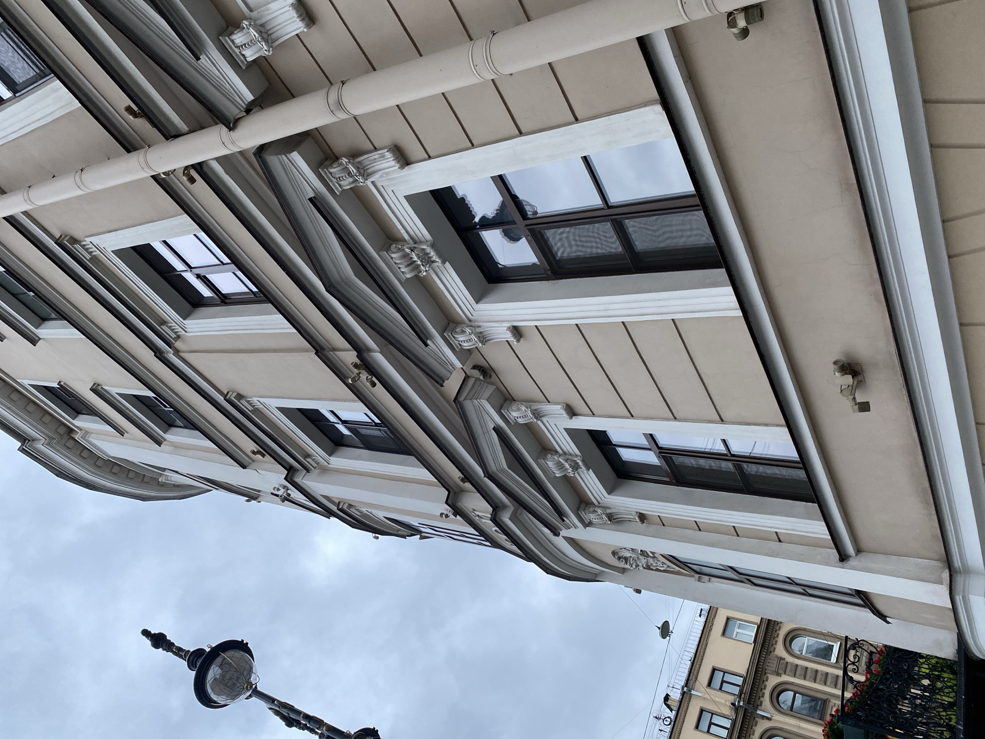 A low-angle shot depicts the facade of a building in Saint Petersburg, Russia, alongside the sky. The architecture showcases a light-colored building with multiple windows, each framed by decorative moldings and corbels. A large rain gutter runs horizontally above the windows.
Below, a portion of a streetlamp is visible against the overcast sky. In the lower right corner, another building is partially seen, constructed with arched windows and decorative details. A black fence partially covers this building.