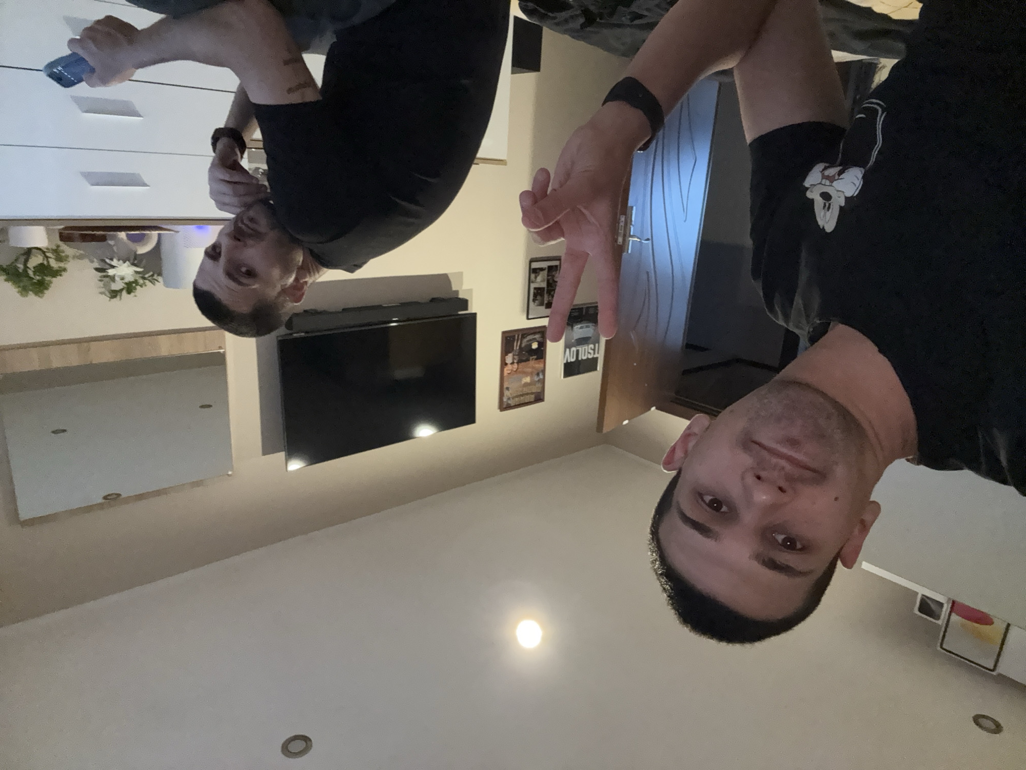 Two adult males are posing for a picture indoors. The man on the right is flashing a peace sign. A television is mounted on the wall behind the men. There are framed pictures on the wall between them and a door. There are recessed lights on the ceiling. A white dresser and a houseplant are visible in the background. The location in the city of Lovech, Bulgaria is unknown.