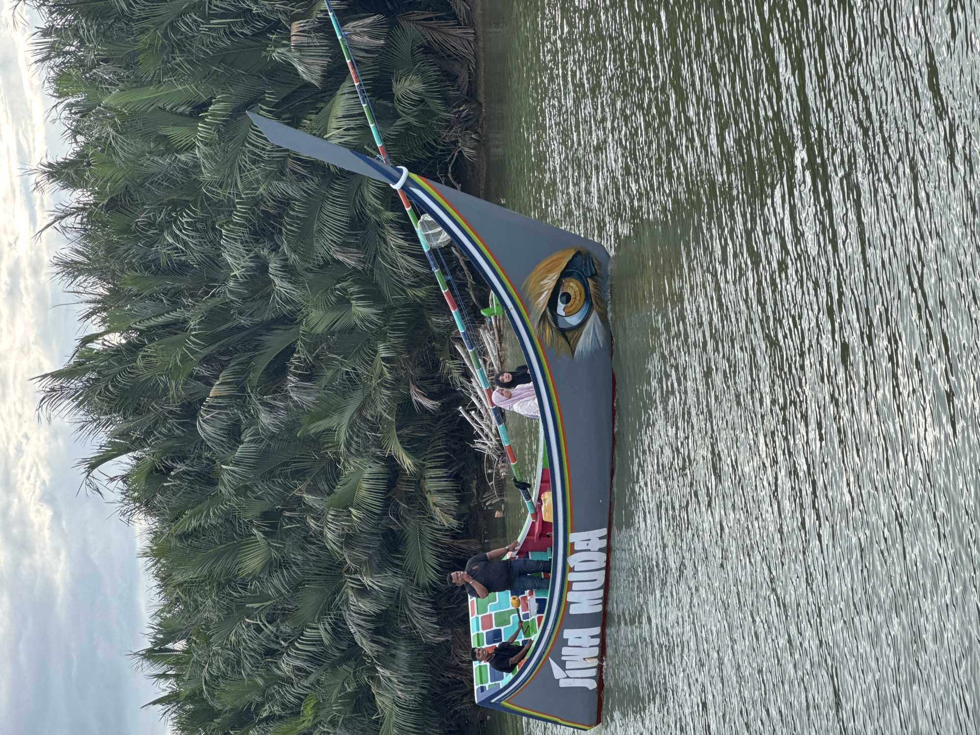 The image shows a boat with a distinctive design, painted with a large eye on its bow, floating on a body of water. The boat is gray with colorful accents along its rim and a vertical, fin-like structure at the front. The words "JIWA MUDA" are prominently displayed on the side of the boat. There are four people on board: two younger males, one of whom is seated and looking forward, and another standing. A man in a black polo shirt and jeans is standing and appears to be gesturing or speaking. A woman in a light-colored hijab is seated towards the back of the boat. The background consists of a dense line of lush green palm trees, suggesting a tropical or subtropical environment. The water is calm with ripples reflecting the overcast sky. The scene is likely located in Reudeuep, Indonesia, as indicated by the prompt.