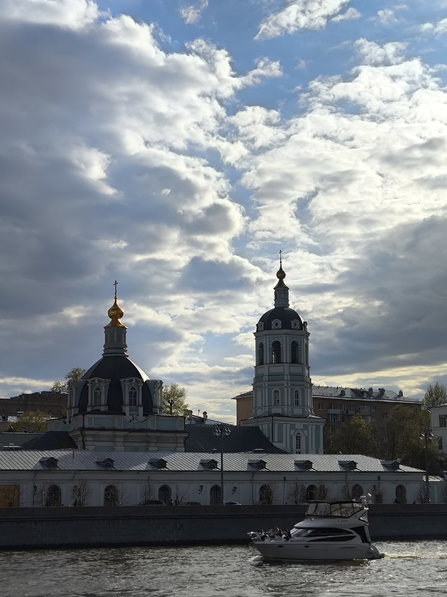 The image captures a cityscape scene, likely in Moscow, Russia, featuring a river, a boat, and a church complex. The foreground is dominated by a body of water, likely a river or a canal. A white motorboat with people on board is seen cruising on the water, creating ripples. Behind the river, a long, low-rise building with a white facade and dark roof can be seen. Architectural details include arched windows and dormers.
To the left, a church complex rises above the low-rise building, featuring two prominent domes. The dome on the left has a gold top, while the one on the right is part of a tall, multi-tiered bell tower. The sky occupies the upper portion of the image and is filled with dramatic clouds, varying in tone from light to dark, and patches of blue sky peek through. Buildings and trees can be seen in the background, indicating an urban environment.