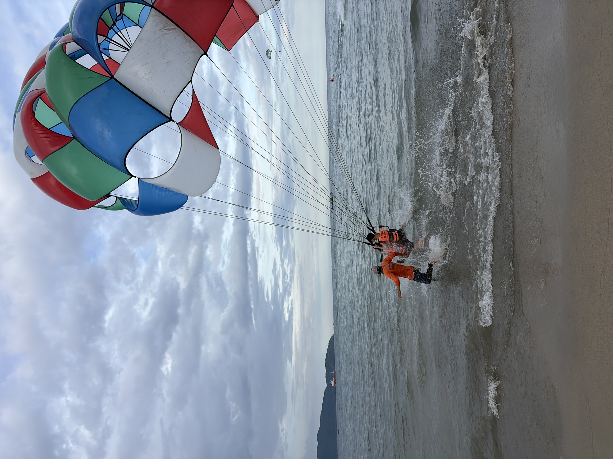 The image depicts a scene at a beach in Đà Nẵng, Vietnam, likely in the vicinity of My Khe Beach, a popular destination for water sports. In the foreground, the edge of a sandy beach meets the ocean. The water is slightly choppy with visible waves breaking near the shore.
Dominating the upper right portion of the image is a large, colorful parachute in shades of red, green, white, and blue, characteristic of parasailing equipment. Several taut lines extend downwards from the parachute.
In the water, close to the shore, two individuals are engaged in parasailing. One person, wearing an orange shirt and a life vest, is running in the shallow water, appearing to assist in launching the parasail. Another person, also wearing a life vest, is seated and attached to the parasailing harness, ready for ascent. The water is splashing around their legs as they move.
In the middle ground, the ocean stretches out towards the horizon under a cloudy, overcast sky. Several other parasailing setups are visible in the distance, with one parachute and two individuals further out at sea. A small boat is also present in the water. On the far left, a large ship is visible in the distance, anchored near a tree-covered mountain range that forms the coastline. The overall impression is a dynamic scene of recreational activity taking place on a somewhat overcast day at a coastal location in Đà Nẵng.