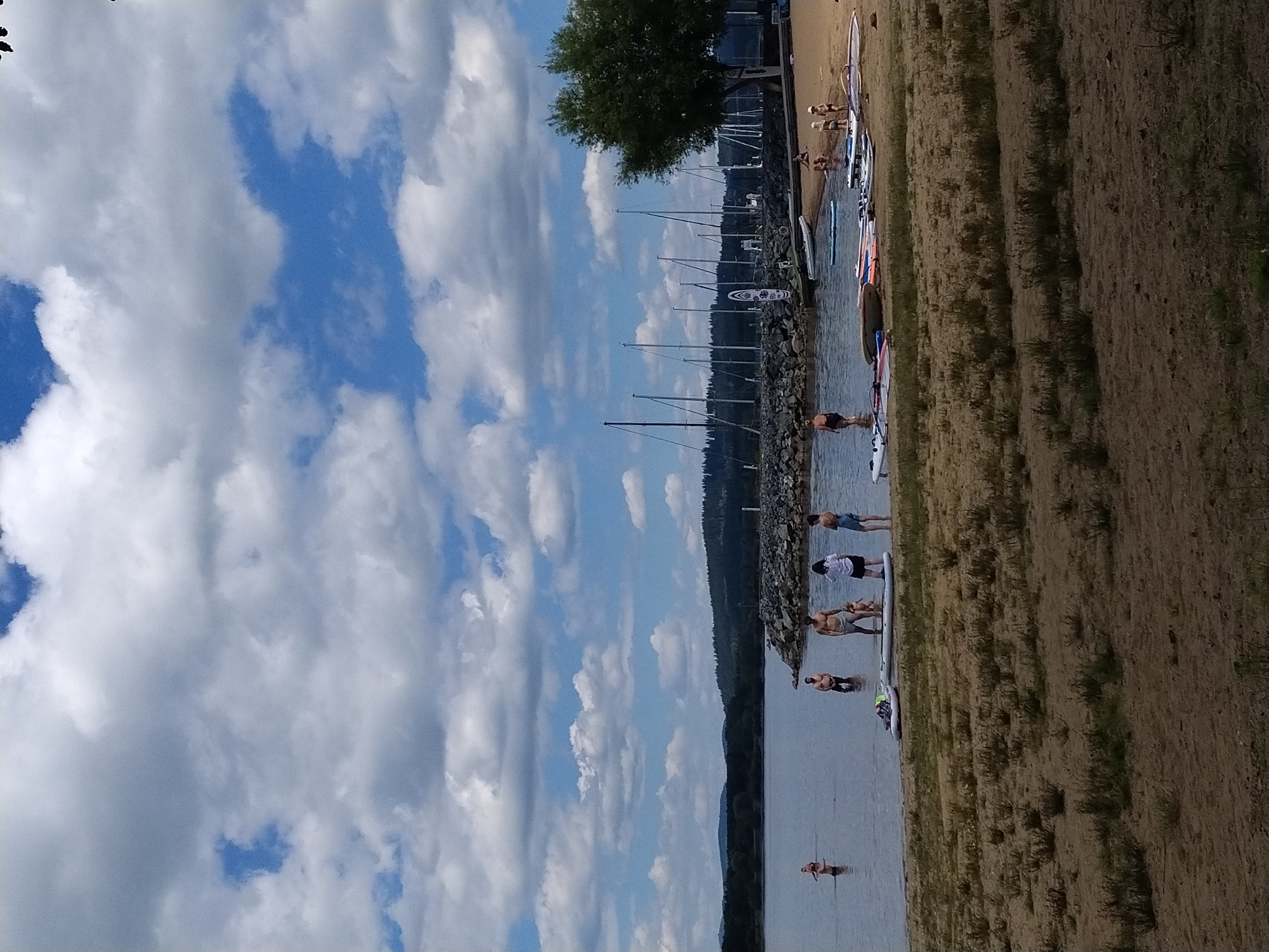 The image depicts a sunny day with scattered clouds over a body of water in Radslav, Czechia. In the foreground, a sandy beach is covered with various surfboards and paddleboards, some of which are partially submerged in the water. Several individuals are present on the beach and in the shallow water. On the beach, a woman wearing a black swimsuit stands near the water's edge, while a group of children plays on the sand. A man and a woman, both in swimwear, are wading in the water, with a young child between them. Further out in the water, a woman stands with her back to the camera. In the midground, a stone breakwater separates the shallow area from a wider expanse of water, where numerous sailboats are docked. The background features a treed shoreline and rolling hills under a bright blue sky with fluffy white clouds.